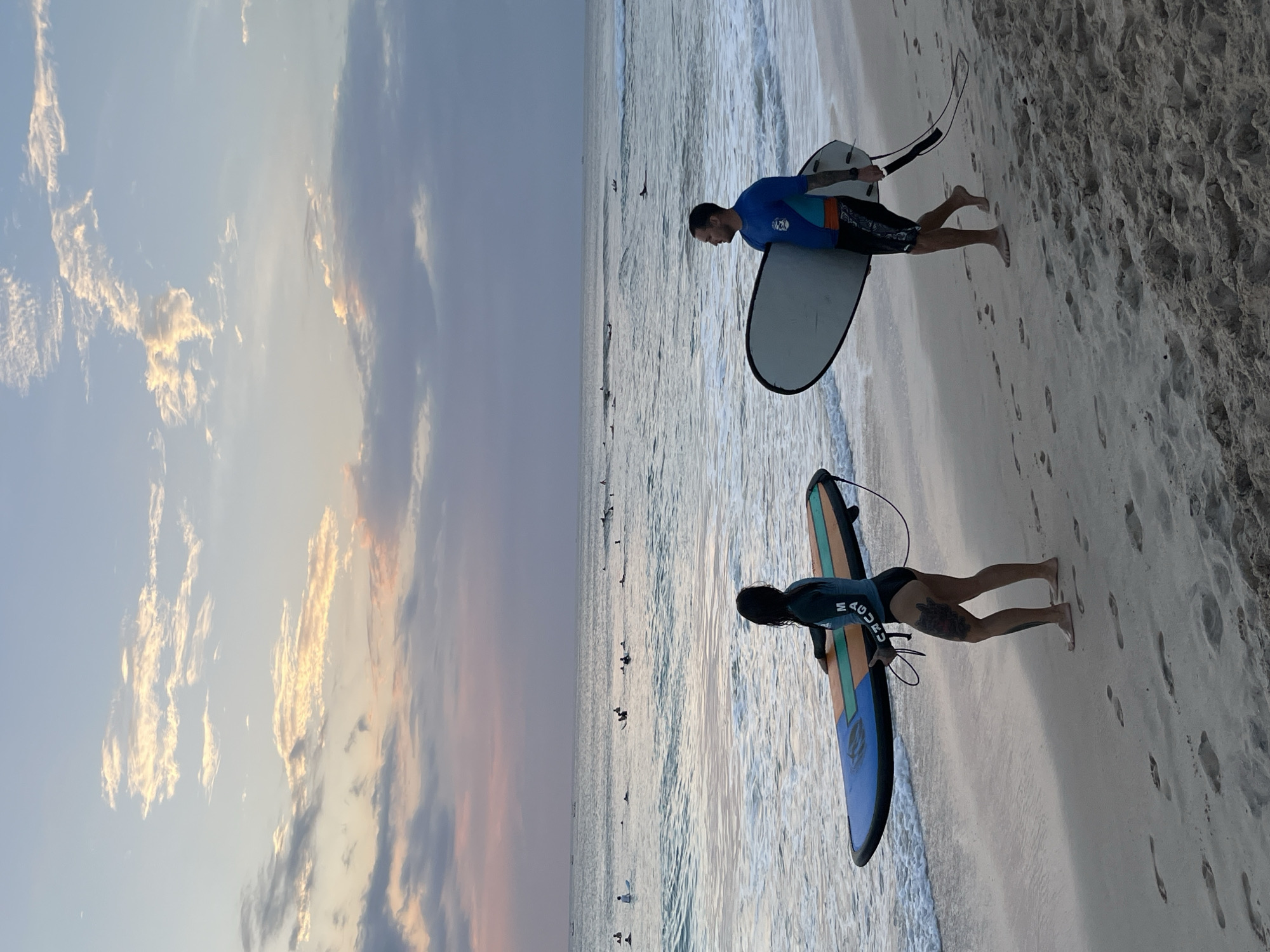 Two surfers, a woman and a man, are walking along the beach in Kuta Selatan, Indonesia, carrying their surfboards. The woman, on the left, wears a dark long-sleeved rash guard and black bikini bottoms. She has tattoos on her legs and is carrying a blue, orange, and yellow surfboard. The man, on the right, wears a blue and turquoise rash guard and patterned shorts. He is carrying a white surfboard. They are walking on a sandy beach with footprints visible. The ocean is to their left, with gentle waves breaking on the shore and several other surfers in the water. The sky above is filled with soft, pastel-colored clouds, suggesting it is either sunrise or sunset. The overall scene depicts a tranquil moment of surfers enjoying the ocean.
