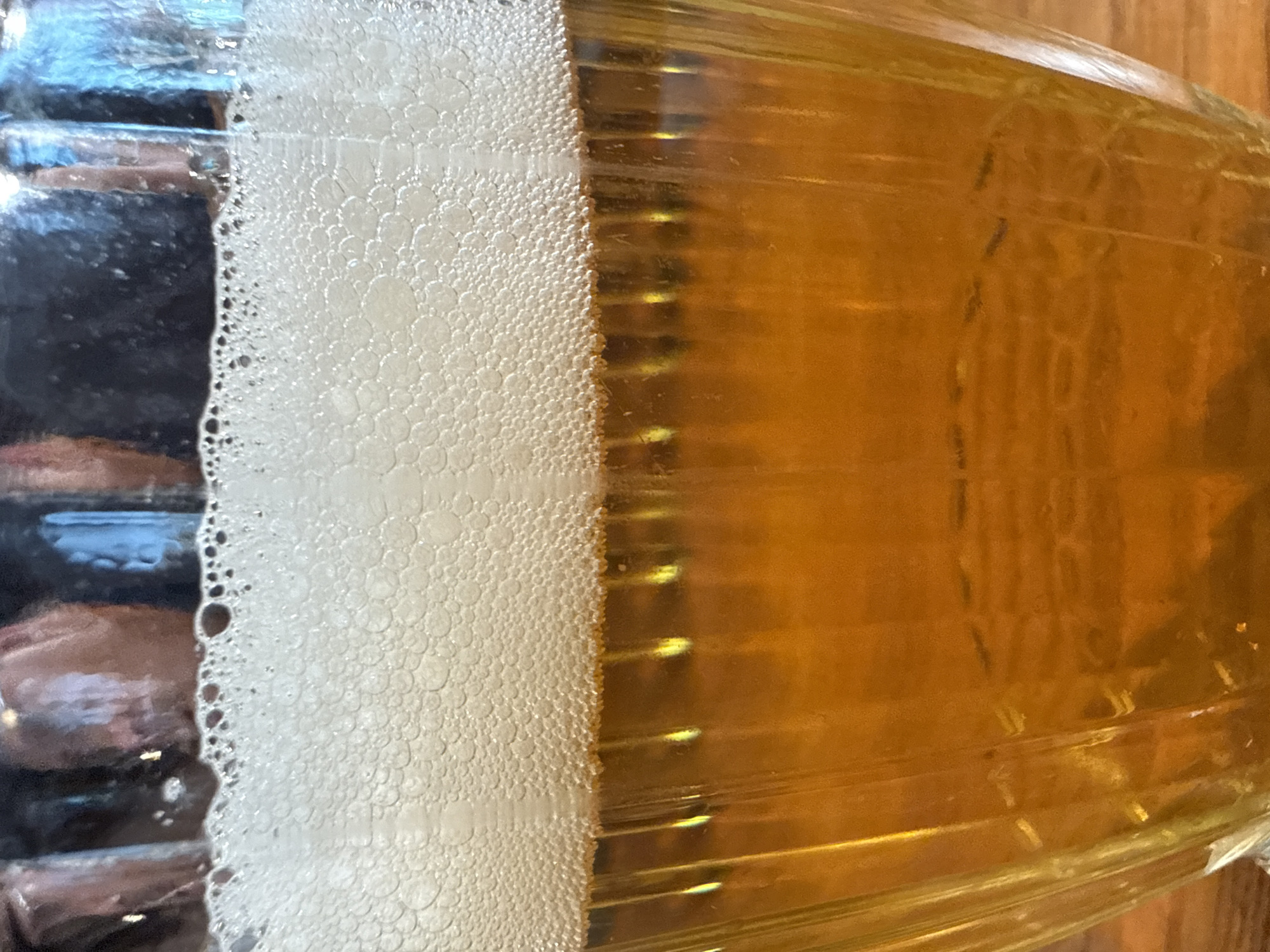 The image is a close-up shot of a glass of beer, viewed from the side. The beer is amber in color and has a thick layer of white foam on top, composed of numerous small bubbles. The glass itself appears to be ribbed or fluted, with vertical lines visible on its surface, refracting the light and creating streaks of yellow and orange. The background is out of focus and shows dark, indistinct shapes which could be reflections or objects behind the glass. The lighting is warm and suggests an indoor setting, possibly a bar or restaurant. There is no discernible indication of the city of Samara, Russia, in this image.