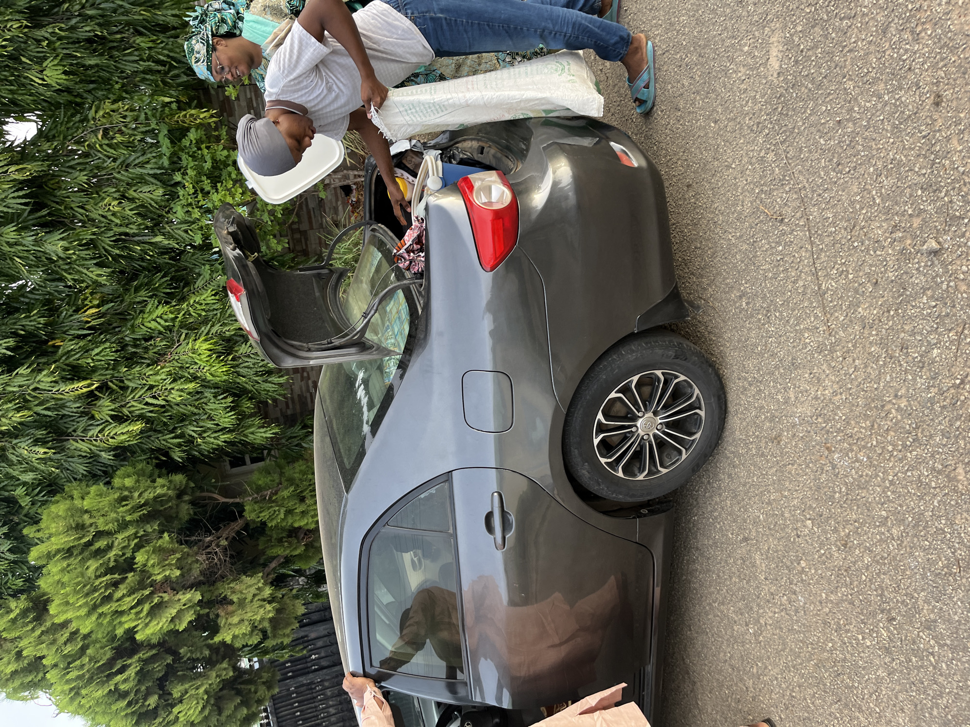 Two individuals are loading items into the open trunk of a dark grey sedan on a paved street. One person wears jeans and a white shirt, holding a white bag above the trunk. The other, wearing a grey head covering, appears to be placing items inside. A white tub rests on the trunk edge. The car's door is ajar, revealing a partial view of the interior. Lush green trees line the roadside. The location is inferred to be in Abuja, Nigeria.