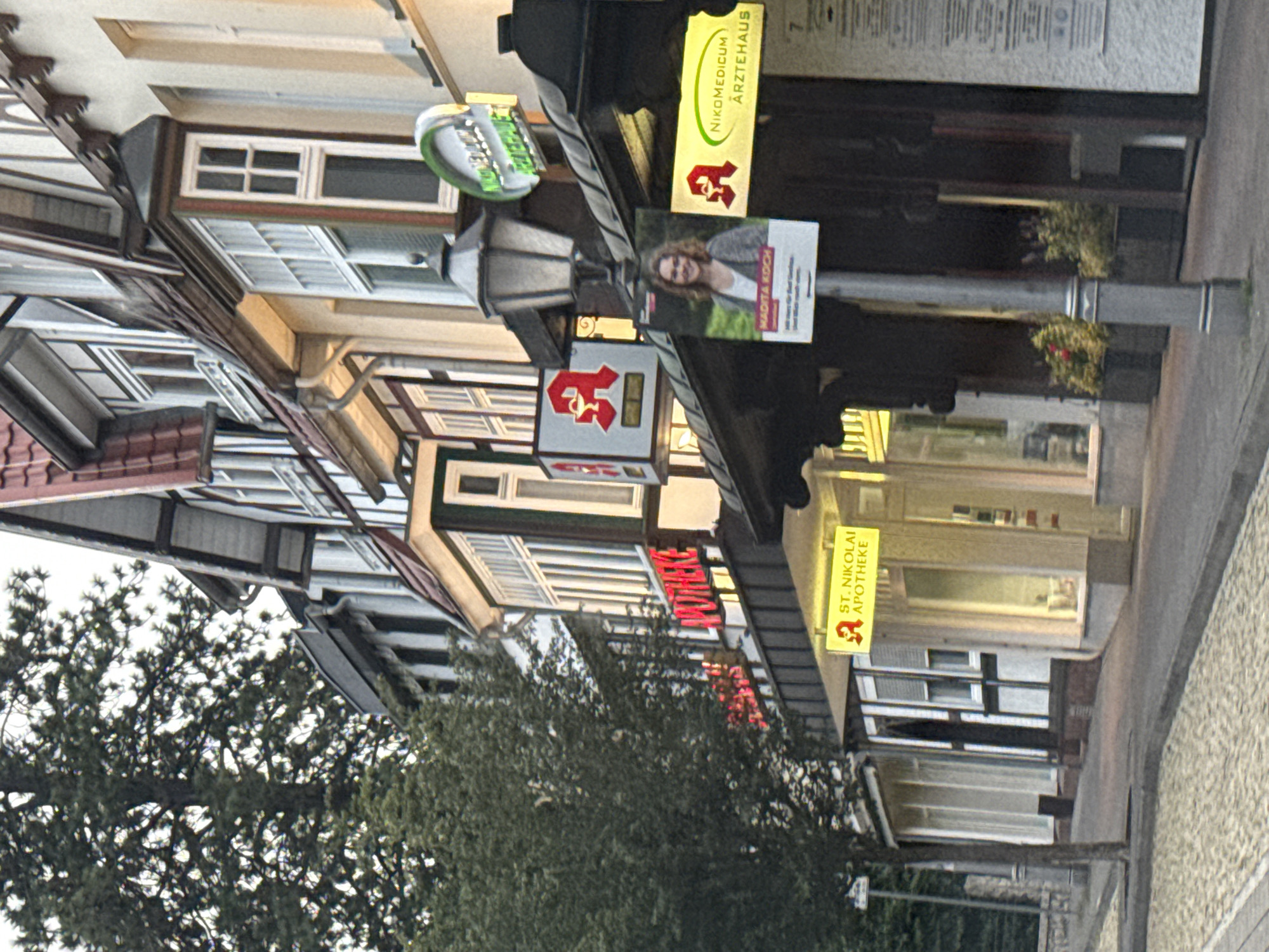 The image shows a street view in Bad Sachsa, Germany, featuring traditional German architecture. Several storefronts are visible, including an "APOTHEKE" (pharmacy) with a prominent red logo featuring a mortar and pestle. Another pharmacy, "ST. NIKOLAI APOTHEKE," is also identifiable by its yellow sign. A lamppost stands in front of one of the shops, with a poster of a woman named "MADITA KOCH" attached to it. The buildings have timber-framed upper stories and tiled roofs. A tree with dense foliage is situated to the left of the frame. The street in the foreground is paved with cobblestones and asphalt. The overall impression is that of a quaint European town street.