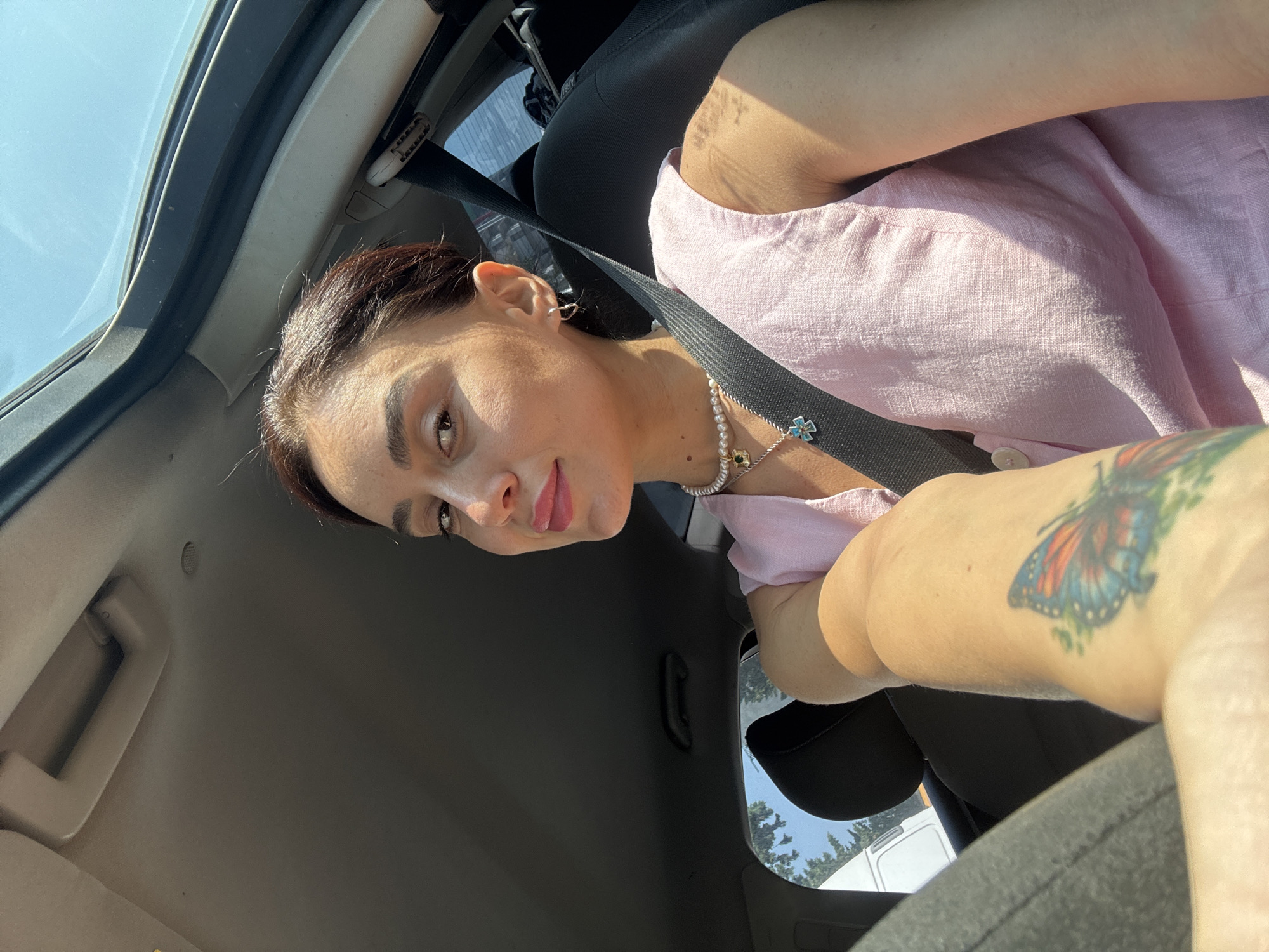 The image shows a woman sitting in the driver's seat of a car, looking towards the camera. She has dark hair pulled back, dark eyes, and is wearing light pink clothing and a pearl necklace. A seatbelt is visible across her chest. A butterfly tattoo is visible on her left arm. The interior of the car, including the dashboard and ceiling, is visible. Outside the car, trees and the sky are visible, suggesting an outdoor setting. The image was taken in Tbilisi, Georgia.