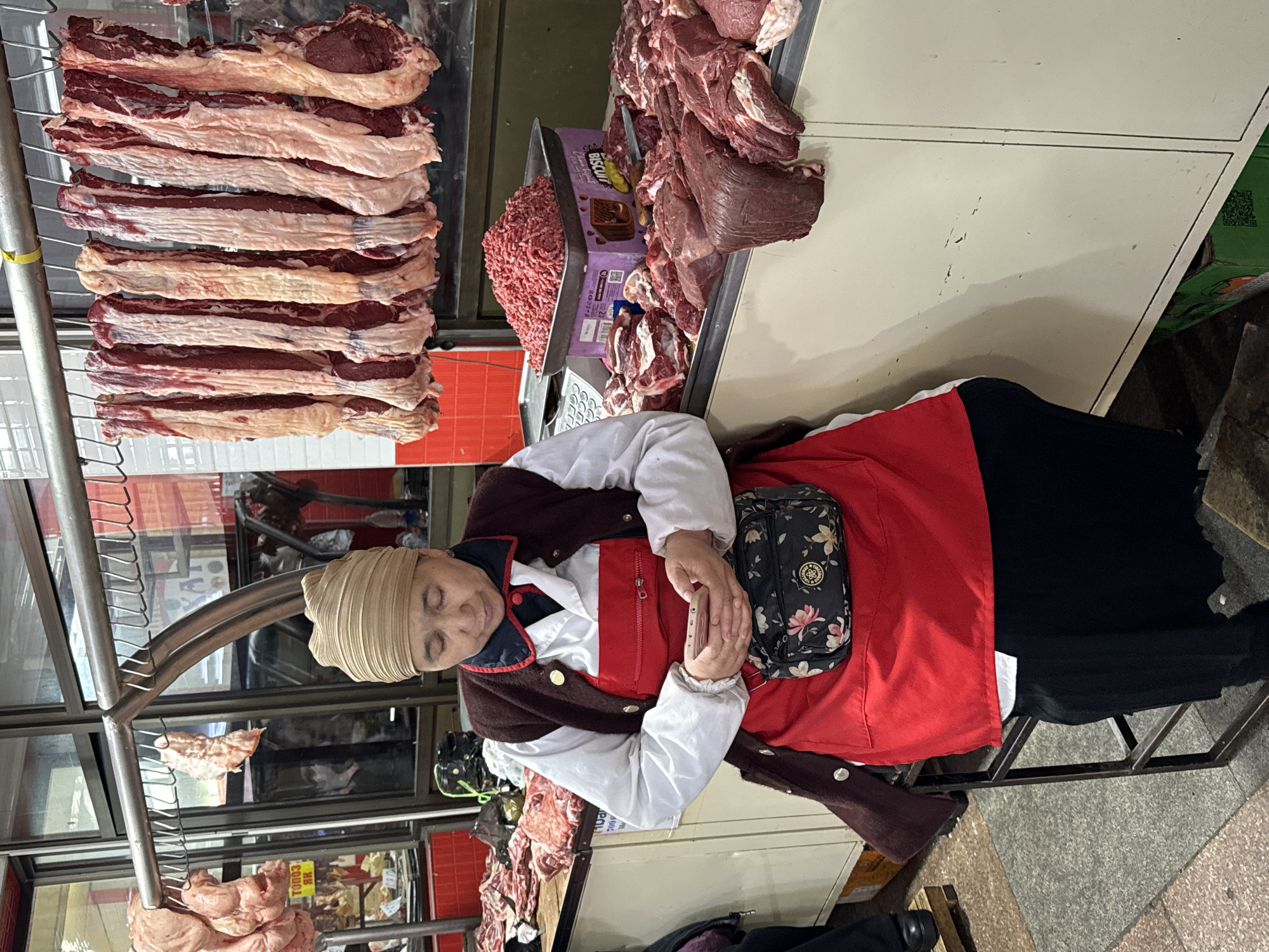 The image captures a bustling scene within an indoor market in Bishkek City, Kyrgyzstan, focusing on a butcher stall. An older woman, likely the vendor, is seated behind a light-colored counter, her eyes closed or downcast, possibly resting or looking at a smartphone held in her hands. She wears a light-colored, pleated head covering, a white long-sleeved shirt, a red vest or apron, and a dark brown cardigan. A small, floral-patterned fanny pack or cross-body bag rests on her lap.
The stall prominently displays various cuts of raw meat. To the left, several large, boneless strips of red meat, possibly beef or lamb, with visible fat, hang vertically from metal hooks on a multi-tiered rack. On the counter to the right, large piles of freshly cut red meat are arranged, along with a tray containing a mound of finely ground meat. A purple box, possibly for sweets or biscuits, labeled "BISCUIL" is visible behind the ground meat, next to the larger meat piles.
The background shows reflections in glass panels, hinting at other market activities or stalls. Metal structures, part of the stall's framework, are visible throughout. On a sign in the background, partially obscured by the metal rack, Cyrillic text, including "ТОНОЗ ЯК" (TONOZ YAK) and other characters, is visible, along with a QR code on a green box. The flooring is a grey tiled or concrete surface. The scene portrays a typical market environment, with the vendor taking a moment of rest amidst her wares.