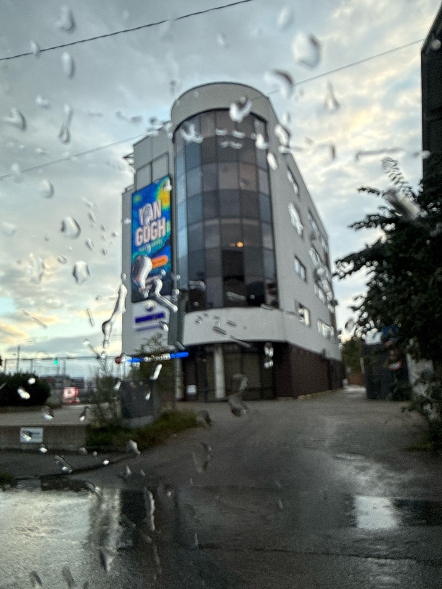 The image depicts a street scene in Tallinn, Estonia, on a cloudy day with visible raindrops on the lens. A modern, multi-story building dominates the center of the frame, featuring a curved glass facade and white exterior walls. A large digital billboard on the side of the building displays "VAN GOGH" in blue and yellow letters.
The street in front of the building appears wet, indicating recent rain. To the left, a street sign and some greenery are visible, along with what looks like traffic lights in the distance. To the right of the building, a dark-colored structure and trees line the street. The overall atmosphere is overcast and slightly blurred, likely due to the rain and the focus of the camera. No people are clearly discernible in the image.