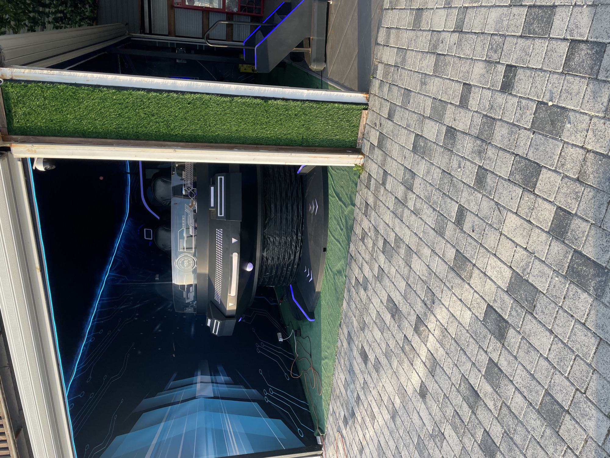 The scene depicts a partially open pavilion or display area in Olginka, Russia. The floor immediately outside is paved with gray concrete pavers in a herringbone pattern. A strip of artificial turf borders the edge of the display area. Inside the structure, there is a futuristic-looking setup featuring illuminated panels with a circuit board design and a large, dark cylindrical object, possibly a display unit or prop. Stairs with blue LED lighting are visible in the background. Electrical cables and wires are scattered on the ground inside the display area. The perspective suggests the photo was taken outdoors.