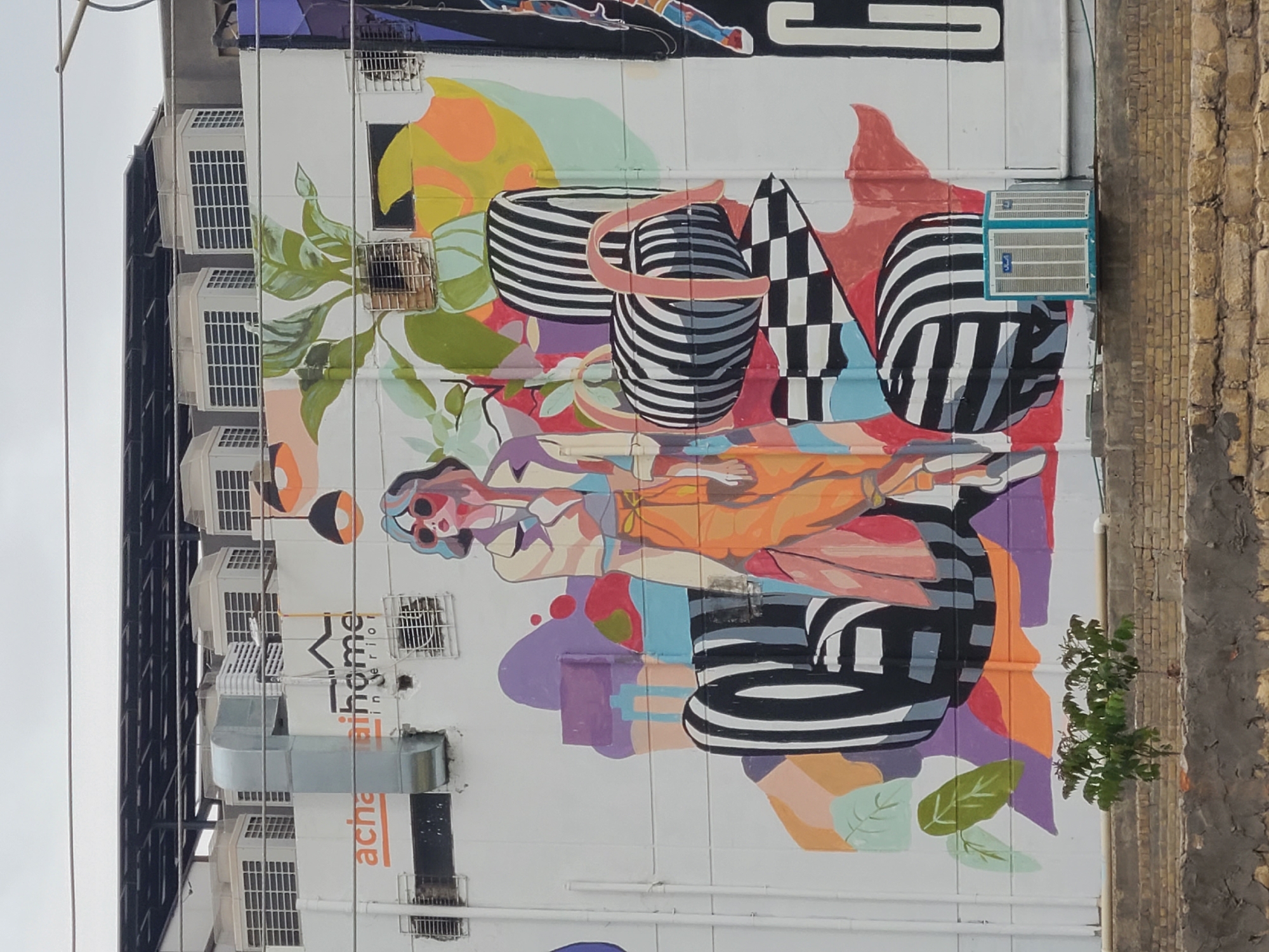 The image shows a mural on a building in Quetta City Tehsil, Pakistan. The mural features abstract shapes and figures, including striped containers, a checkerboard pattern, and floral elements. The colors are vibrant and include orange, purple, green, and blue. The building also has air conditioning units installed on the facade. A small tree grows next to the building, near the edge of the mural.