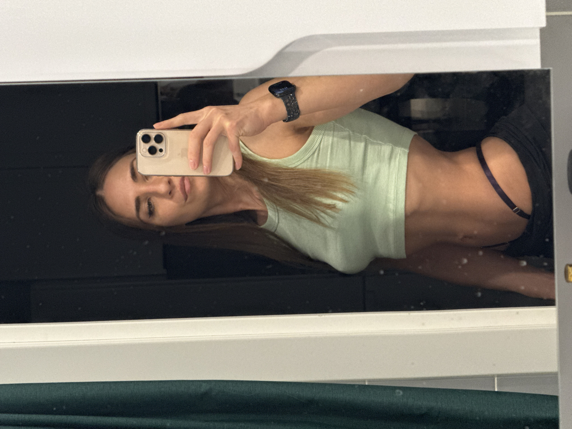 A woman takes a selfie in a mirror. She wears a light green crop top and black bottoms with a blue elastic band visible. She holds a smartphone with a beige case. A smartwatch is on her wrist. The mirror reflects a dark room or space. The background is indistinct, likely due to the mirror's reflection and lighting. The location is Lyubertsy, Russia. There is no way to prove this statement to be true or false based on the information provided.