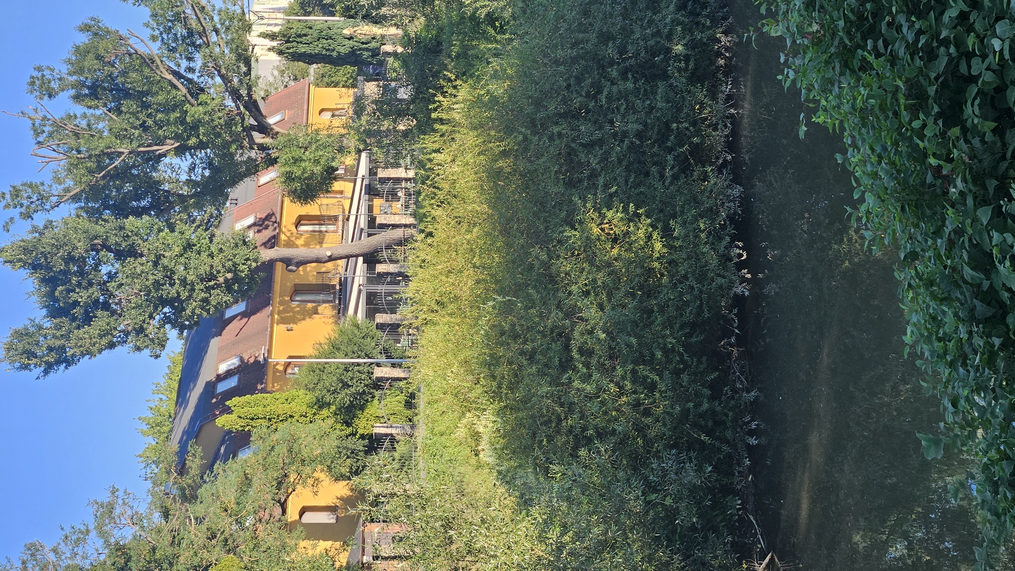 The image depicts a residential building with a yellow facade and a red tiled roof, situated behind a dense thicket of green trees and bushes. The building features several dormer windows on the roof and balconies on the second floor, suggesting it is a multi-story dwelling or a hotel. A dark, calm body of water, likely a river or a canal, occupies the foreground, reflecting the surrounding vegetation and the building. The scene is bathed in the warm, golden light of what appears to be either early morning or late afternoon. The overall impression is one of a serene, natural setting within a developed area.
While the specific location within Prešov, Slovakia, cannot be definitively determined from the visual information alone, the architectural style of the building and the presence of lush vegetation are consistent with many areas in the city. Prešov, being the third-largest city in Slovakia, has a mix of urban and natural landscapes, with rivers and parks often bordering residential and commercial areas. The image captures a common scene found in such environments.