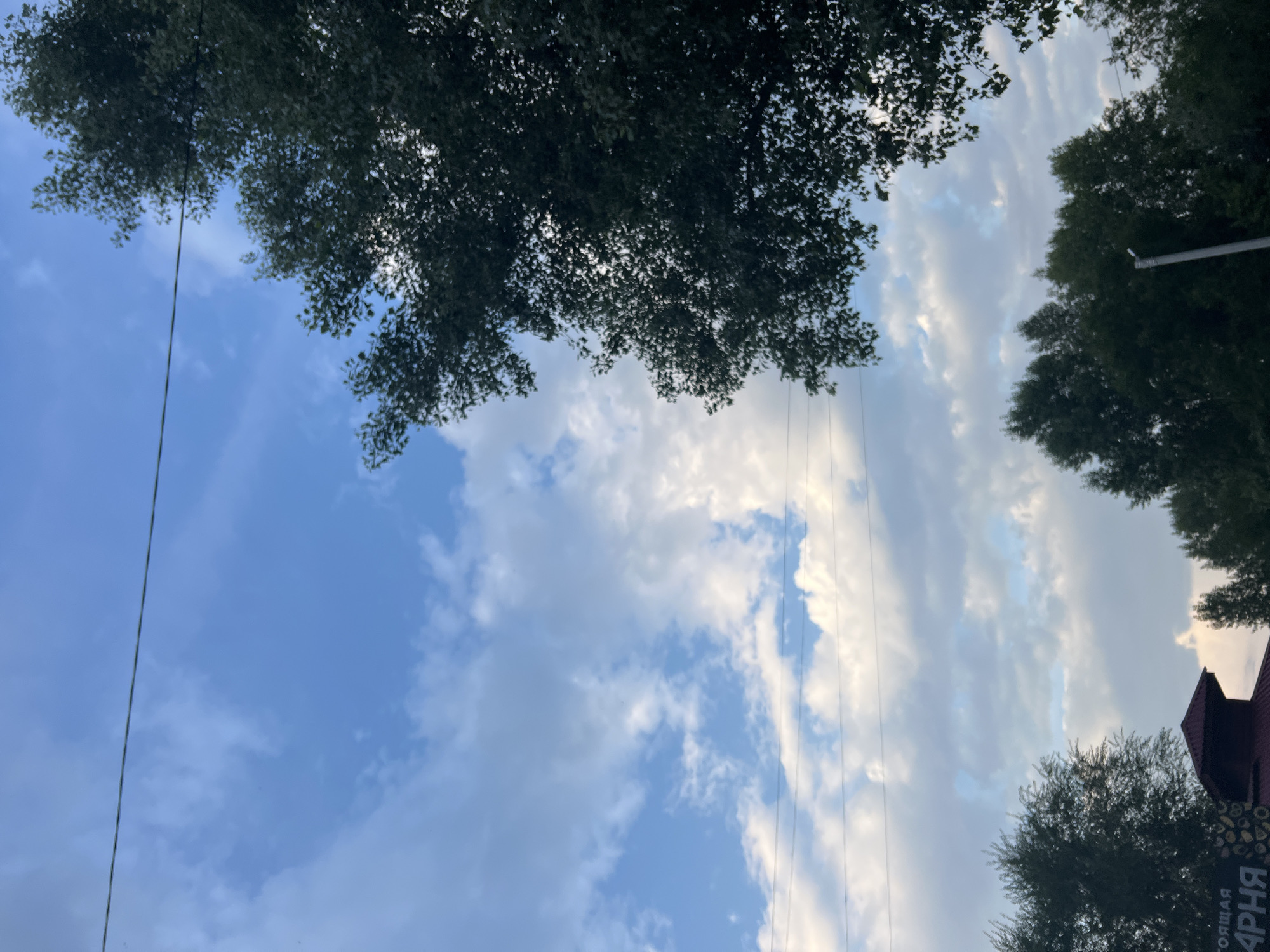 A wide-angle, low-angle outdoor shot captures a section of the sky with fluffy white clouds against a blue background. A thin black power line stretches horizontally across the upper portion of the frame. On the right side, the dark green foliage of a mature tree dominates the upper right quadrant, its leaves casting dappled shadows. In the lower portion of the image, more trees are visible, silhouetted against the sky. A dark red, gabled roof of a building is partially visible in the lower left, with some text on a sign to the left of the roof. A grey pole with a small white fixture at the top is visible in the lower right. The overall impression is of an outdoor scene, possibly in Ulyanovsk, Russia, on a partially cloudy day.