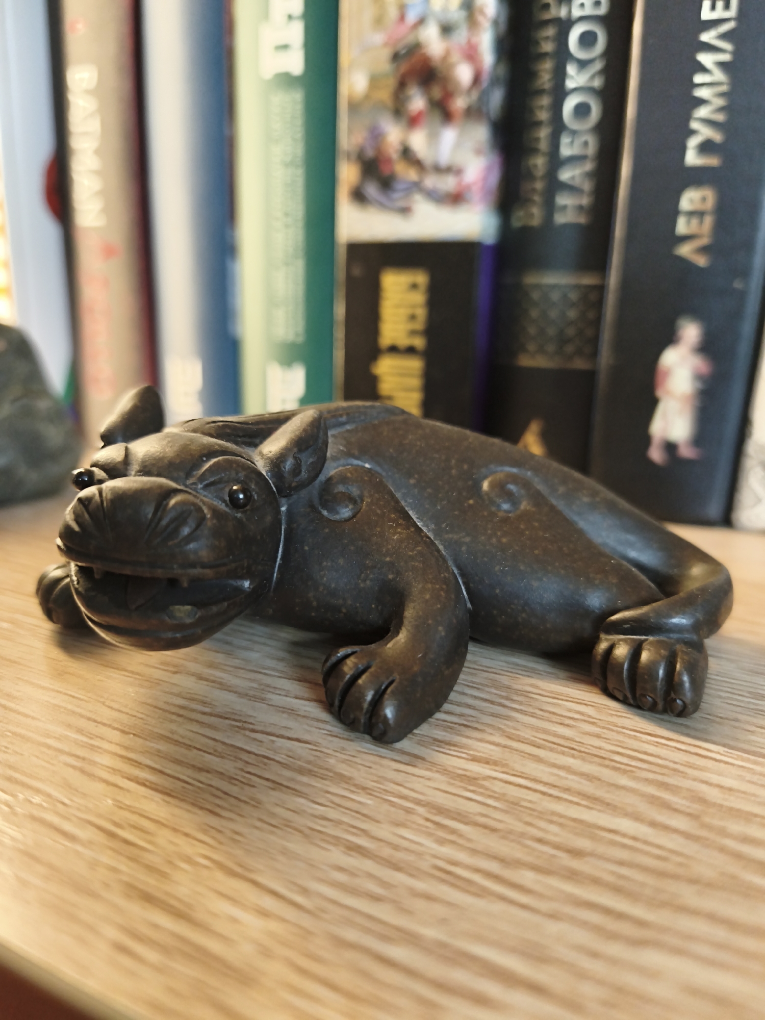 A dark, bronze-colored figurine of a mythical creature, possibly a stylized dog or lion, is positioned on a wooden surface. The figurine is depicted lying down, with its head turned slightly to the left. It has a wide-open mouth with visible teeth, round, dark eyes, and prominent ears. The body features carved details, including a swirl pattern on its back.
In the background, a row of books is visible, with their spines facing the camera. The books are of various colors, including cream, blue, green, and black. Some of the spines have visible text. One book spine on the far left appears to have the word "BATMAN" written vertically in a light color. Another black book spine in the middle displays Cyrillic text. The book on the far right also has Cyrillic text, with the phrase "ЛЕВ ГУМИЛЕВ" clearly legible.
The lighting suggests an indoor setting, likely during the daytime, with light falling from the left, casting shadows from the figurine onto the wooden surface. The wooden surface itself has a distinct grain pattern. The overall scene is a close-up view of the figurine on a shelf, with books in the background. There are no people or animals other than the figurine itself. The context suggests a home or office environment, possibly a display of collectibles. The location context of Astana, Kazakhstan, is provided but not visually evident within the image itself.