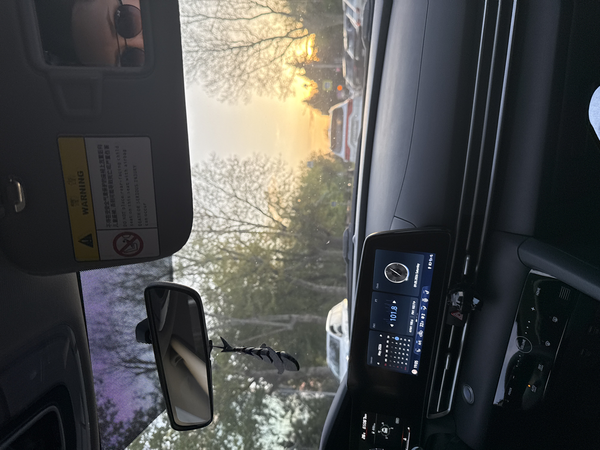 Here is a detailed description of the image:
**Scene:** Interior view of a car, likely parked or stationary. The viewpoint is from the driver's seat.
**Subjects:**
* **Driver:** Reflected in the sun visor mirror, wearing sunglasses.
* **Car Dashboard:** Featuring a digital display showing the radio, clock, and date (May 9, 2026). There are also climate control buttons.
* **Rearview Mirror:** A standard car rearview mirror with a decorative ornament (possibly a shark figurine) hanging from it.
* **Exterior view:** Visible through the windshield, a street scene with trees, a sunset or sunrise, and other vehicles.
**Objects:**
* **Sun Visor:** Displaying a warning label.
* **Digital Display:** Shows radio, clock, and date information.
* **Rearview Mirror Ornament:** Shark figurine.
* **Warning Label:** Attached to the sun visor.
**Environment:**
* **Exterior:** The outside scene appears to be a city street with trees lining the road. The lighting suggests early morning or late afternoon due to the low sun angle.
* **Interior:** Modern car interior with a focus on the dashboard and driver's area.