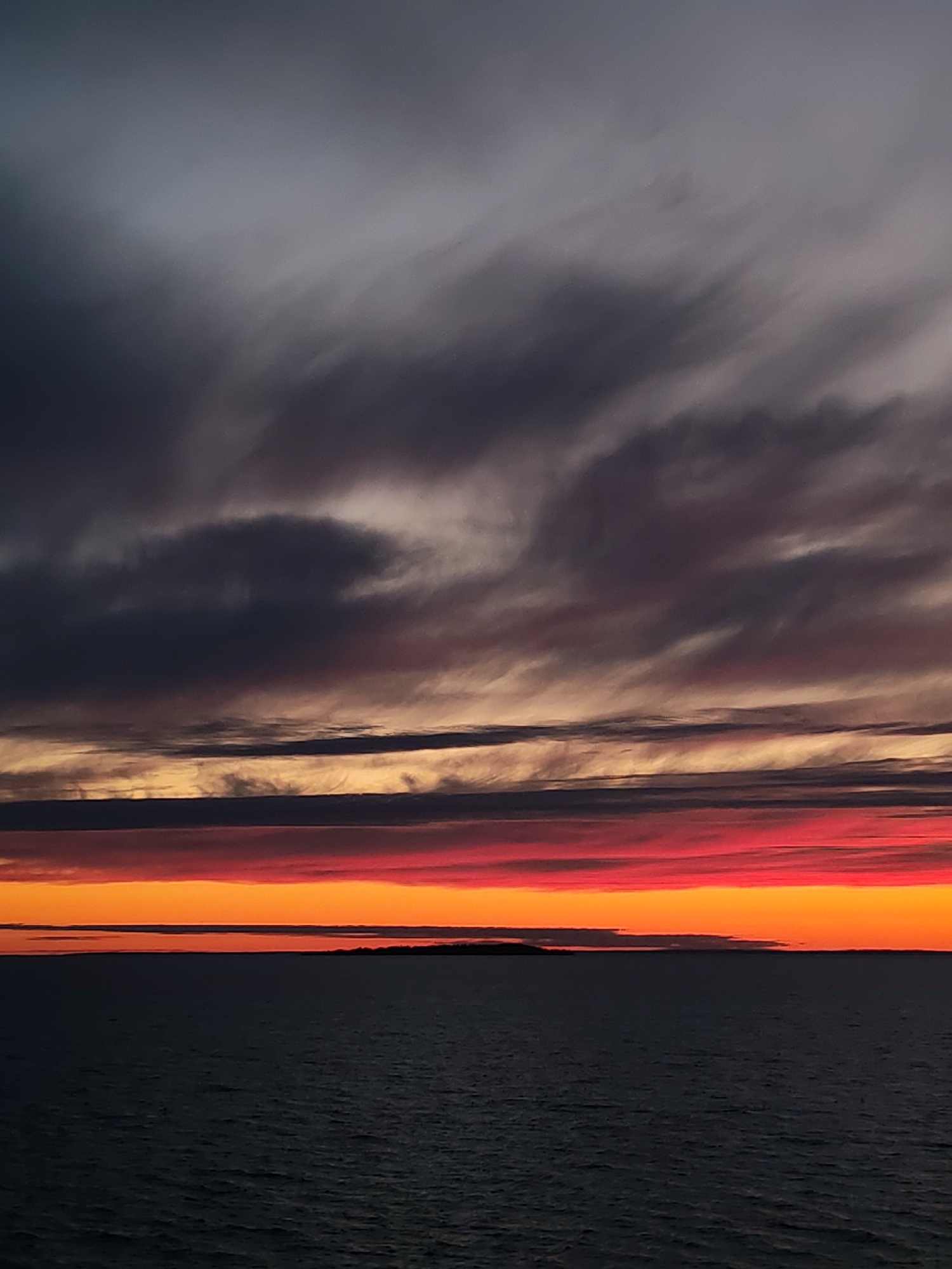 The image depicts a dramatic sunset over a large body of water, likely a sea or lake, in an unknown region of Russia. The sky dominates the frame, showcasing multiple layers of clouds painted with vivid sunset colors.
In the upper portion of the sky, dark, heavy clouds dominate, appearing in shades of grey, dark blue, and muted purple, with some areas showing subtle wispy textures. Moving downwards, the clouds transition into progressively warmer and more intense hues. A distinct band of deep reds and oranges stretches horizontally across the mid-sky, forming vibrant, layered strips interspersed with darker cloud formations. Closer to the horizon, the sky glows with a brilliant band of fiery orange and red, indicating the immediate presence of the setting sun's light. The contrast between the dark, heavy clouds above and the luminous, colorful layers below creates a profound sense of depth and drama.
Below the colorful sky, a vast expanse of calm, dark water extends into the foreground. The water's surface is a deep, uniform blue-grey, with subtle textures suggesting gentle ripples, and reflects very little of the sky's intense colors. On the distant horizon, a faint, dark landmass or island is visible, creating a thin line between the water and the lowest band of light.
The scene evokes a sense of serenity and natural grandeur, characteristic of dusk or late evening. There are no people, objects (apart from the natural landscape), or visible text in the image. The dramatic lighting and color palette strongly indicate that the photograph was taken during sunset.