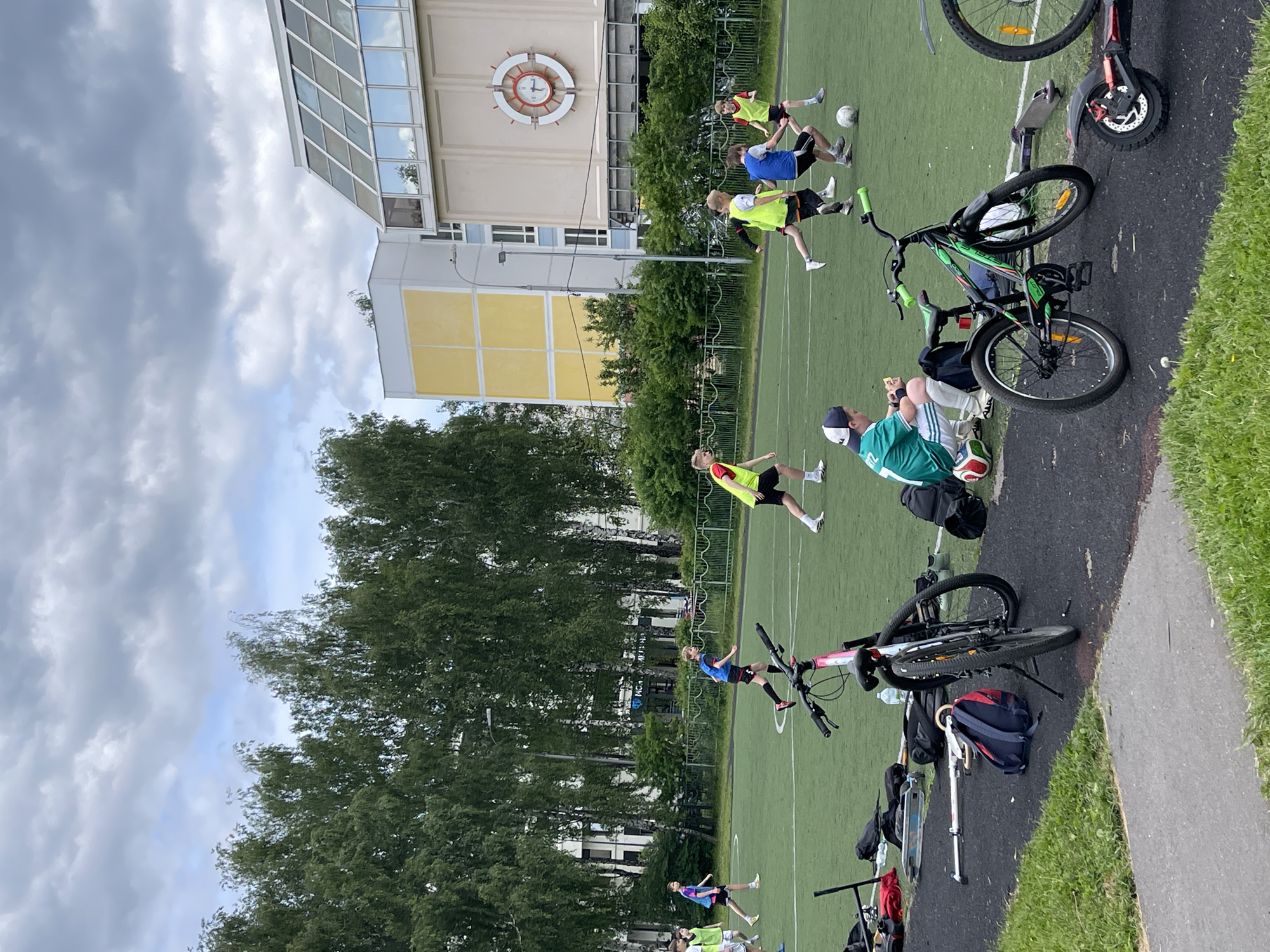 A group of young people are playing soccer on a green artificial turf field next to a building with a clock face on its facade. Several bicycles and scooters are parked on the adjacent paved area. Trees are visible in the background, and the sky is cloudy. It is Moscow, Russia.
