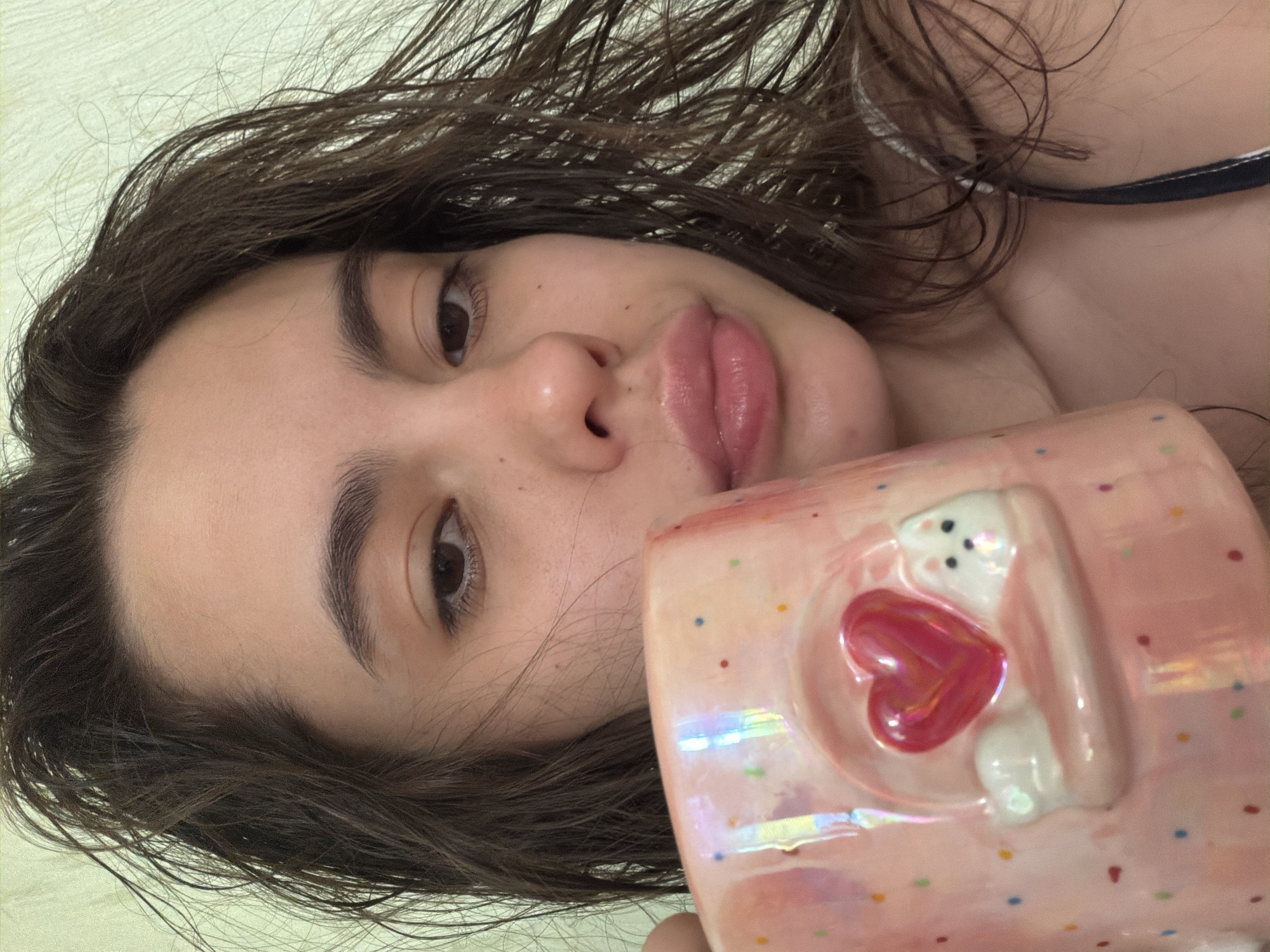 A close-up selfie shows a young woman with wet, dark brown hair and light skin. She has full lips and dark eyebrows. She holds a pink mug with a white rabbit design and a red heart in front of her face. The background appears to be a light-colored wall. The woman is the primary subject.
There is no verifiable evidence to confirm that the picture was taken in the city of Chaikovsky, Russia.