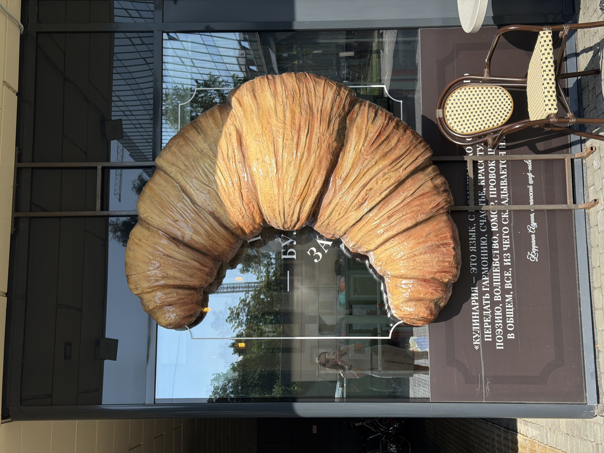 This is a vertical, eye-level, outdoor shot of a large, stylized croissant sculpture displayed on the glass facade of a building. The croissant is a warm, golden brown, with visible folds and a slightly glossy texture, suggesting it's made of a material like plastic or painted plaster. It is positioned prominently in the center of the frame, arching over text on the building's exterior.
Behind the croissant, the glass reflects the environment. We see a grid-like structure overhead, possibly part of the building's architecture or a covered walkway. Trees with green foliage are visible, suggesting it's daytime and likely spring or summer. Sunlight casts shadows on the facade, indicating a bright day.
To the right of the croissant, a brown wicker chair with a woven seat and back is visible, suggesting an outdoor seating area, perhaps for a cafe or restaurant.
Text is visible on the dark brown facade below and to the right of the croissant. The text appears to be in Russian. Part of it reads: «КУЛИНАРИЯ — ЭТО ЯЗЫК, С ПЕРЕДАТЬ ГАРМОНИЮ, СЧАСЬЕ, КРАСОТУ, ПОЭЗИЮ, ВОЛШЕБСТВО, ЮМОР, ПРОВОК... В ОБЩЕМ, ВСЕ, ИЗ ЧЕГО СК. АДЫВАЕТСЯ Н». Another line reads "Буугана Адриа, чанский шеф-пов". These phrases translate roughly to "Culinary is a language, to convey harmony, happiness, beauty, poetry, magic, humor, provocation... In general, everything that is needed." and "Buggana Adria, a Siberian chef."
In the reflection on the glass, a person is visible taking a photo of the croissant with their phone, and another person is partially visible in the background. The overall impression is of a charming, perhaps slightly whimsical, storefront advertising a bakery or cafe. The location context suggests this is Saint Petersburg, Russia.