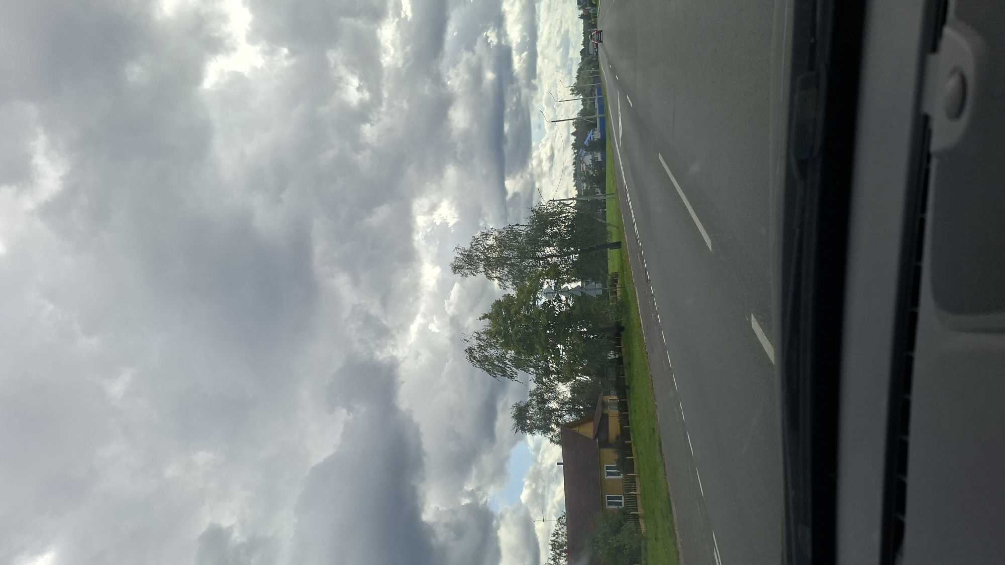 The image captures a cloudy day scene from inside a vehicle, looking out towards a rural road and residential area. A grey, overcast sky dominates the upper portion of the frame. On the left side of the road, a yellow house with a dark red roof is visible, bordered by a wooden fence and green grass. Several tall trees, appearing to be birch trees due to their slender trunks and leafy canopies, stand between the house and the road. The road itself is paved with asphalt and marked with white dashed lines, indicating lanes. In the distance, along the right side of the road, streetlights and more houses with blue roofs can be seen. A red car is driving away in the distance on the road. The foreground shows the dashboard and windshield of the vehicle. The scene is likely located on a road leading into or out of Myadzyel, Belarus, given the typical rural landscape and architecture.