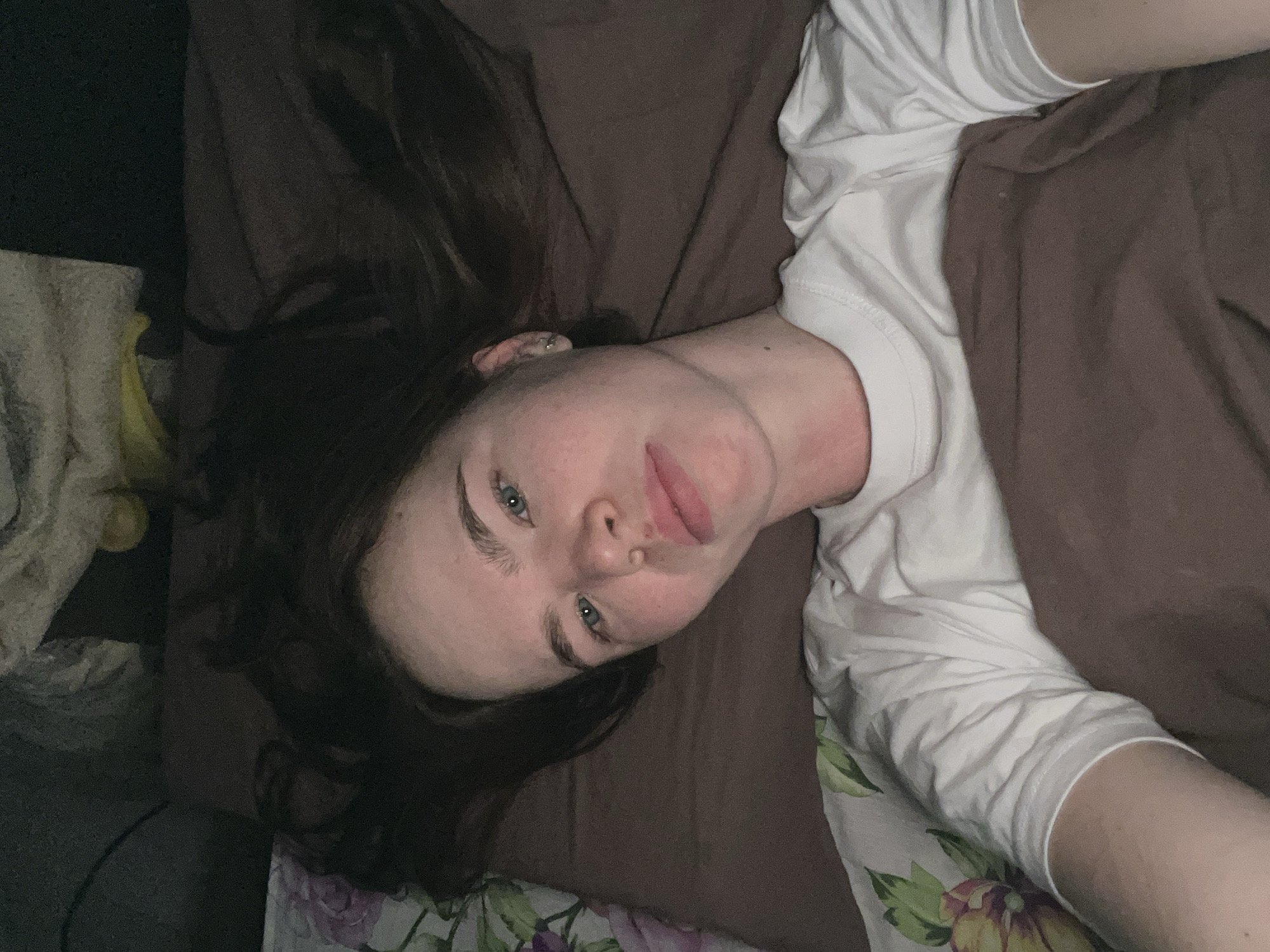 A young woman lies on a bed in what appears to be a dimly lit room. She has fair skin, dark hair, and blue eyes. Her head is tilted, and she is looking directly at the camera. She is wearing a white t-shirt. A brown blanket covers part of her body. Floral patterned sheets are visible beneath the blanket. The location is not identifiable as Chaikovsky, Russia, from the visual data provided.