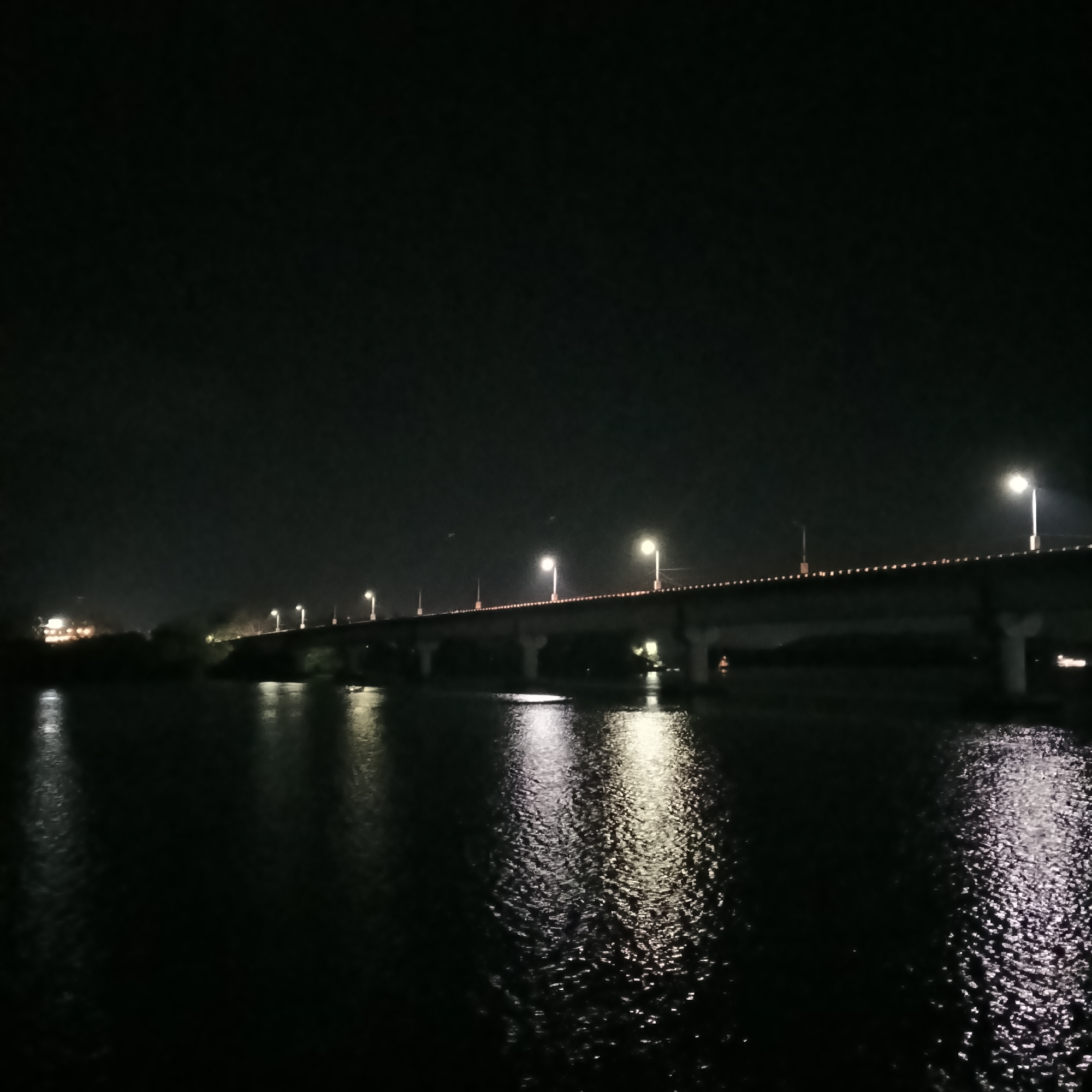 The image depicts a bridge at night, spanning a body of water. The bridge is illuminated by evenly spaced streetlights. The water reflects the light from the bridge, creating shimmering patterns. The sky is dark and clear. Vegetation is visible on the far bank of the water. The location is Oxel, India.