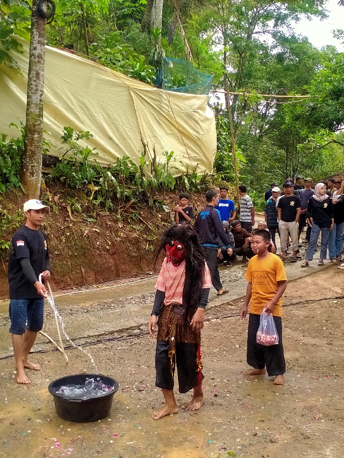 In Gempolan, Indonesia, an outdoor scene depicts a group of individuals engaged in an activity. In the foreground, a barefoot man wearing a white cap and dark t-shirt holds a white hose, directing a stream of water into a black plastic basin containing water and scattered pink/red objects. Adjacent to him, a barefoot figure in a red, grotesque mask with long, dark, matted hair stands, wearing a pink top, a brown patterned lower garment, and dark pants. This figure's lower right pant leg is discolored red. To the right, a barefoot boy in an orange t-shirt holds a clear plastic bag containing small red objects. The ground is wet, muddy dirt with some paved sections, and scattered with red/pink petals. In the midground, a crowd of approximately 15-20 individuals, including men, women (one wearing a hijab), and children, observe from behind the main figures. Behind them, a steep, heavily vegetated hillside features a large, light-yellow tarpaulin covering a structure, with some green netting visible above. Lush green trees and foliage form the background.