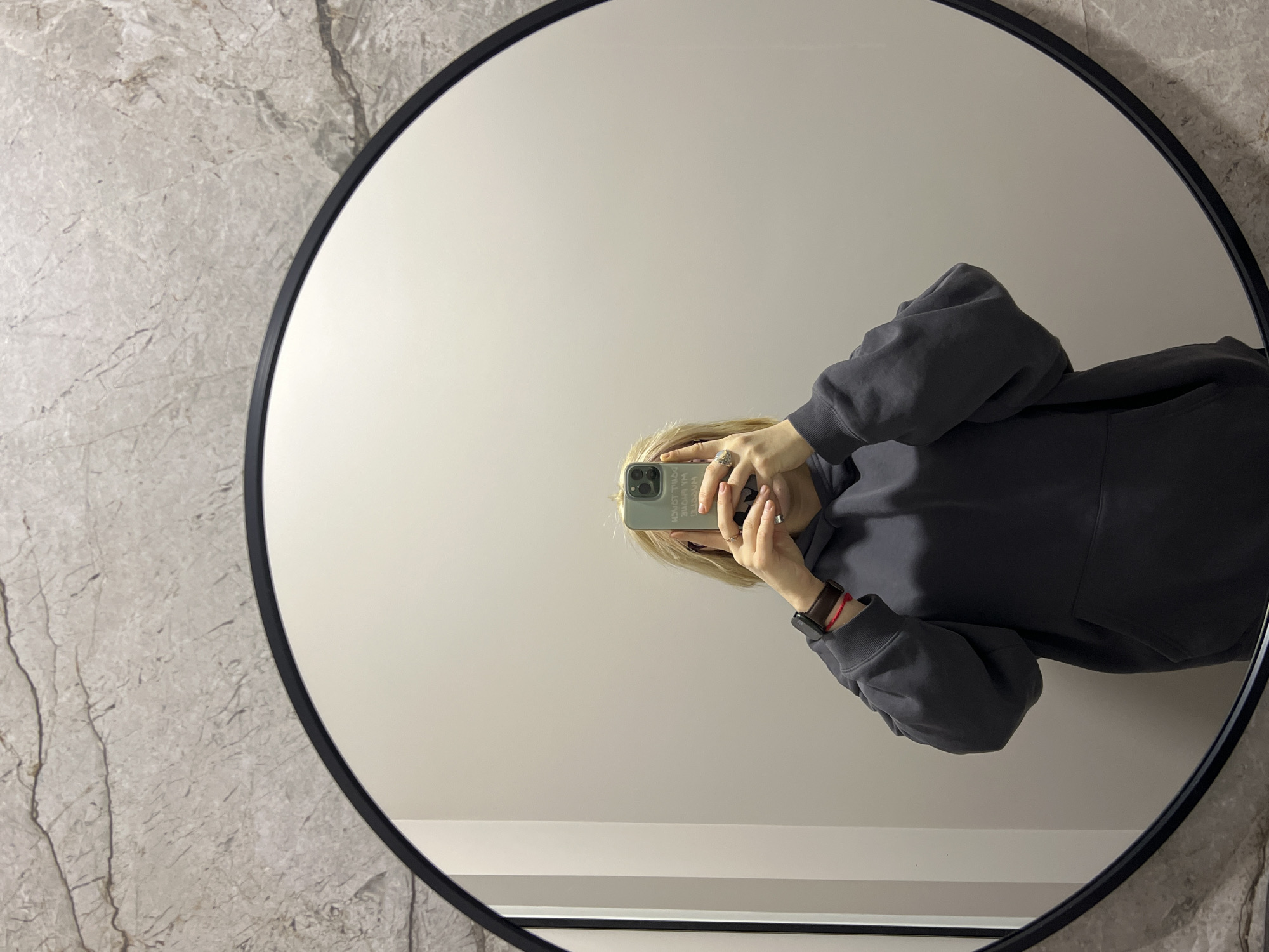 A person is reflected in a round mirror with a black frame. The mirror is mounted on a wall that has a marble-like texture on the left and a solid light color in the center. The person is holding a phone with a green case, obscuring their face in the reflection. They are wearing a gray hoodie and a watch. The location cannot be determined specifically as Odesa, Ukraine with the given information.