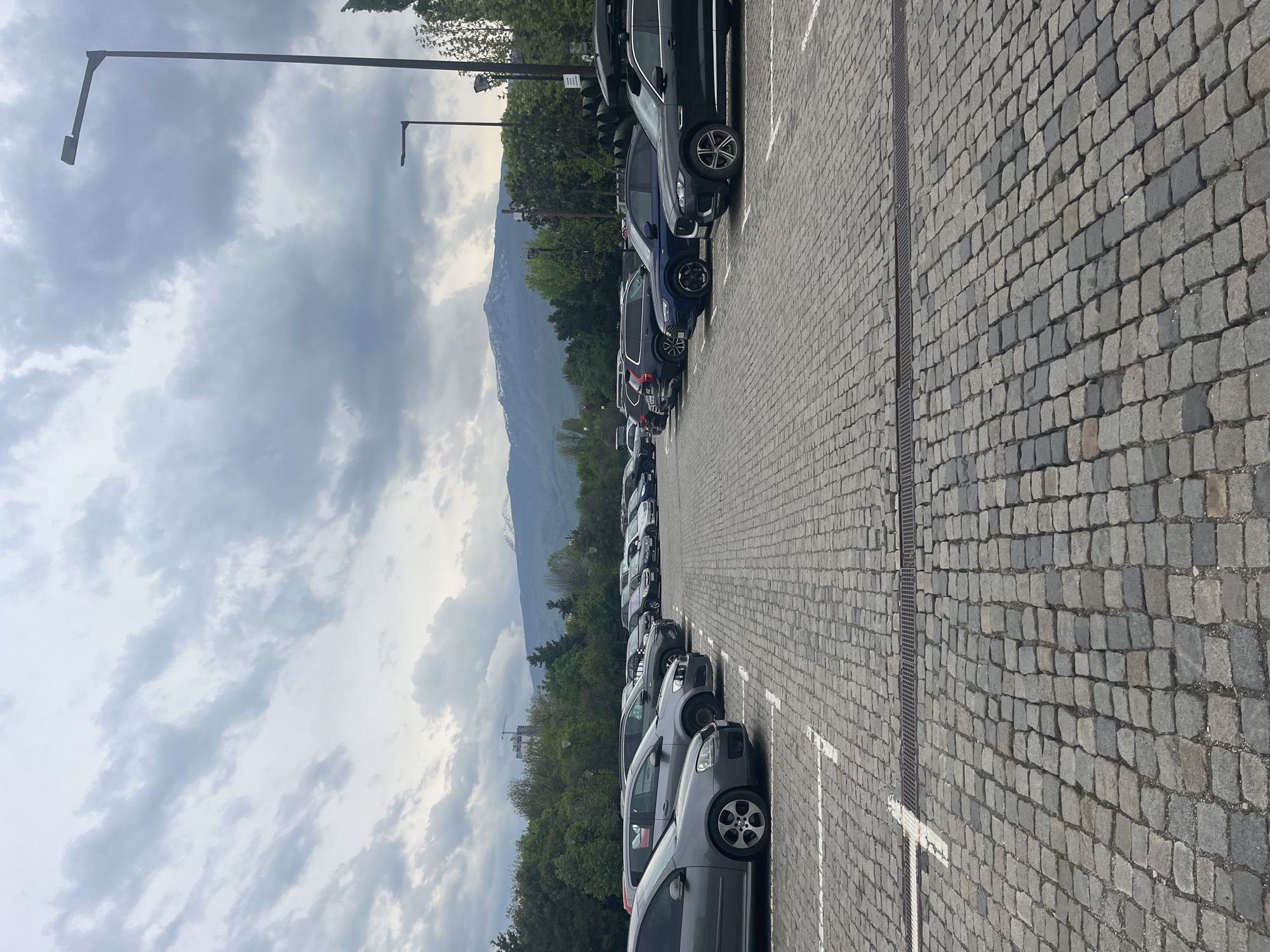 The image captures an outdoor parking lot under an overcast sky, with mountains visible in the distant background. The parking area is paved with grey cobblestones, arranged in a rectilinear pattern, and marked with white painted lines for individual parking spaces. A narrow metal drain grate runs across the cobblestone surface in the mid-ground, paralleling the parking rows.
Numerous cars are parked in two main rows, facing away from the viewer. The vehicles are primarily sedans and SUVs, varying in model and color, with many appearing in shades of grey, black, and dark blue. Details such as alloy wheels are visible on several cars.
Beyond the parking lot, a band of green trees and lush foliage stretches across the mid-ground, separating the man-made infrastructure from the natural landscape. A faint construction crane can be seen rising above the trees on the left side of the frame. In the far background, majestic mountains dominate the horizon, characterized by rugged slopes and hints of lighter, possibly snow-covered or rocky, patches near their peaks.
The sky is largely covered with a mix of grey and white clouds, indicating an overcast or partly cloudy day. Two dark metal streetlights, featuring long, angled arms, extend into the upper portion of the frame against the sky.
The scene depicts a typical daytime setting, with no people visible. While some small stickers or tags are present on cars, no decipherable text can be read. The overall impression is a mundane parking facility situated in a scenic location, consistent with a mountainous region such as Sofia, Bulgaria.