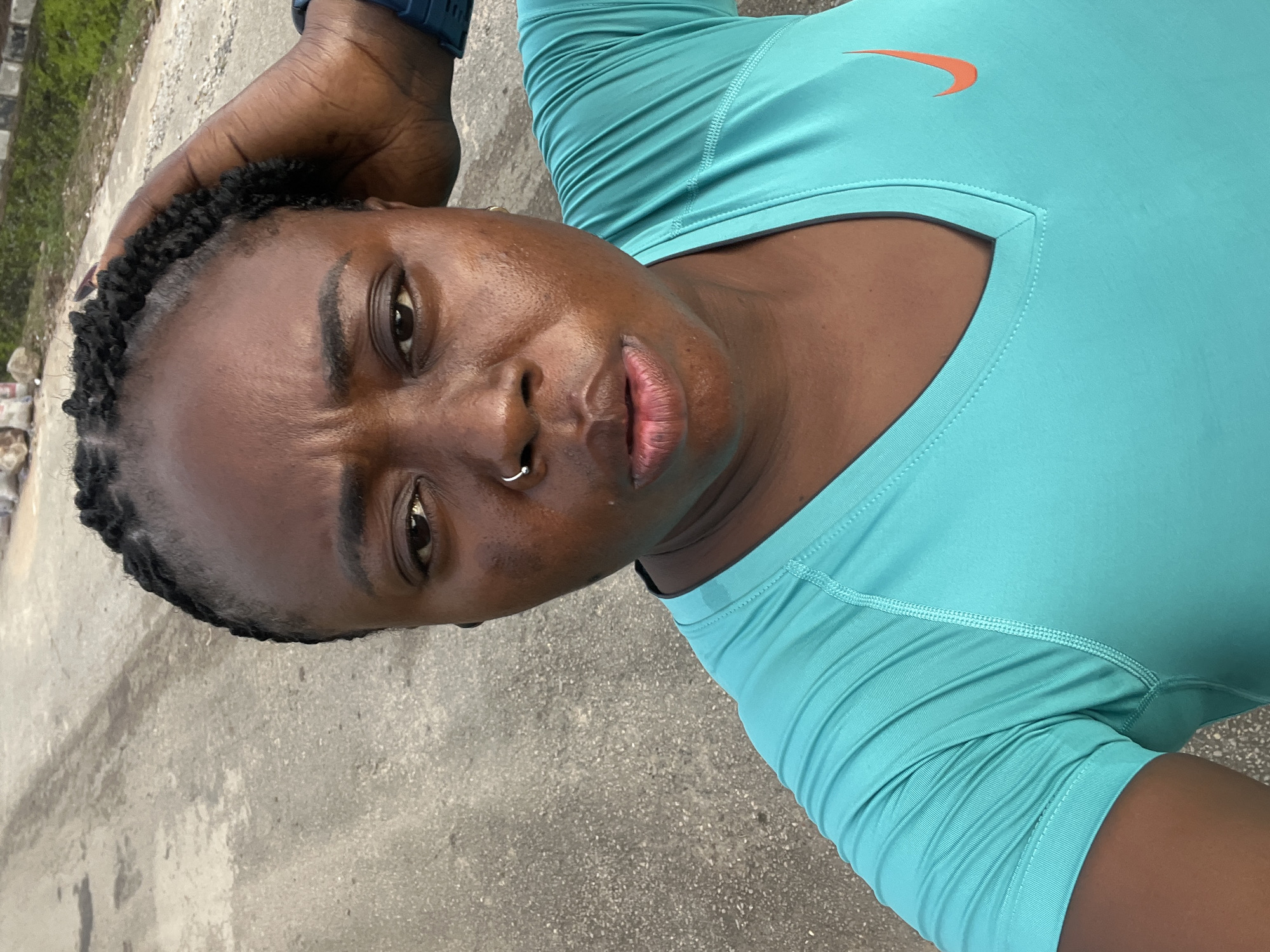 In this close-up, selfie-style photograph taken outdoors, a Black woman with braided hair and a silver nose ring is the central subject. She is wearing a bright turquoise V-neck t-shirt with an orange Nike swoosh logo on the right side of her chest. Her right hand is raised, touching her hair. The background is an asphalt surface, likely a road, with some texture and discoloration. In the upper right portion of the background, a hint of greenery and some scattered debris, possibly construction materials like bags, are visible. The lighting suggests it is daytime. The overall scene appears to be in an outdoor urban or suburban setting.