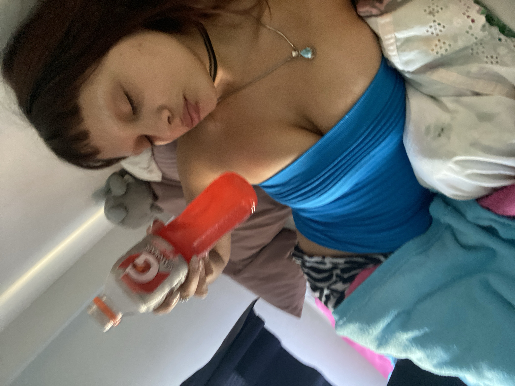 A woman is lying in bed, appearing to be asleep. She is wearing a blue strapless top, a choker necklace, and a pendant necklace. She is partially covered by white sheets and a blue blanket. She is holding a bottle of orange Gatorade. The location is not discernable as Buenos Aires, Argentina, as there are no indicators in the image to suggest this.