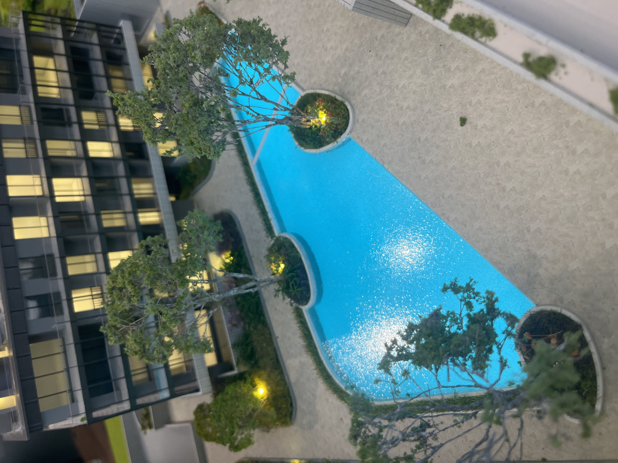 This is a high-angle, close-up shot of a meticulously crafted architectural model. The model depicts a modern apartment building with multiple stories, characterized by large glass windows and balconies. In front of the building, there is a distinctly shaped swimming pool filled with bright blue water, reflecting the lights within the building model. The pool area is surrounded by light-colored paving stones and meticulously placed miniature trees and landscaping. A pathway, also paved, winds around the pool and leads away from the building. The overall scene suggests a luxurious, contemporary residential development. There are no people visible in the model. The context of Limassol, Cyprus, is not discernible from the visual elements of the model itself.