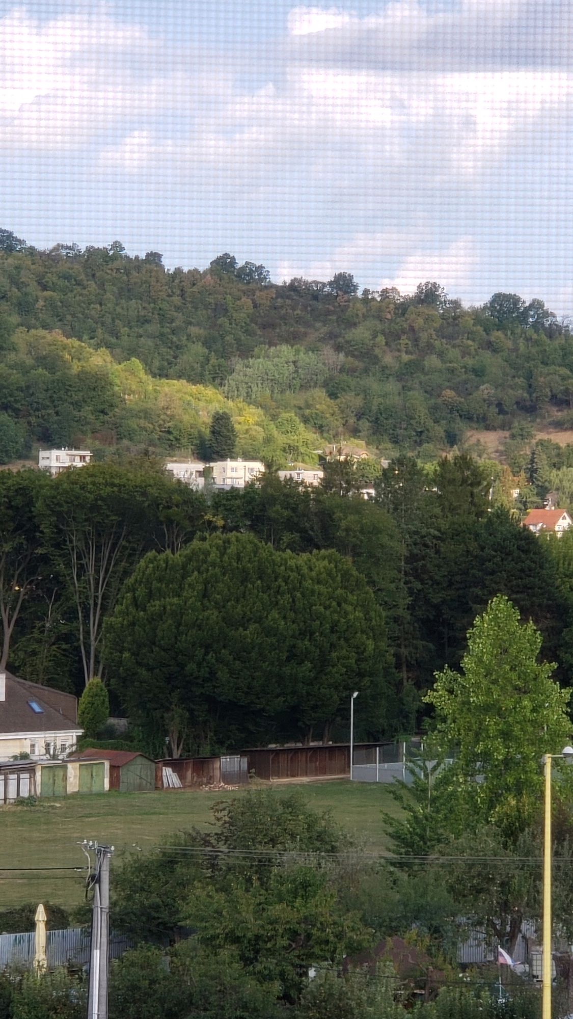 The image shows a suburban landscape with residential buildings nestled into a hillside covered with dense deciduous forest. In the foreground, there is a green lawn with several outbuildings including garages and sheds. A paved area with a fence and a sports court is visible behind the outbuildings. A tall yellow pole, likely a street lamp, stands on the right side of the image. Power lines and utility poles are also present in the foreground. The sky is partly cloudy with a bright, overcast appearance. The overall scene suggests a tranquil, natural setting on the outskirts of a developed area. The architecture of the houses on the hillside suggests a European setting, possibly in the city of Prešov, Slovakia, which is known for its hilly terrain and surrounding forests.