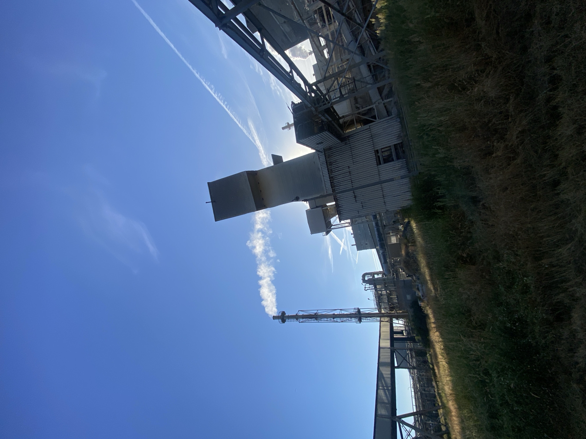 The image depicts an industrial facility under a clear blue sky with scattered contrails. In the foreground, tall dry grass and green vegetation are visible on a slope. To the left, a covered walkway supported by metal beams stretches across the frame, leading towards the industrial structures. In the center-left, a tall, slender smokestack emits a plume of white smoke. To the right of the smokestack, the industrial complex features several large, grey buildings with corrugated metal siding, elevated conveyor belts, and complex pipework. Sunlight filters through the structures, creating shadows and highlighting parts of the buildings. The overall impression is one of an active industrial site with a bright sky. The location is identified as the city of Police, Poland.