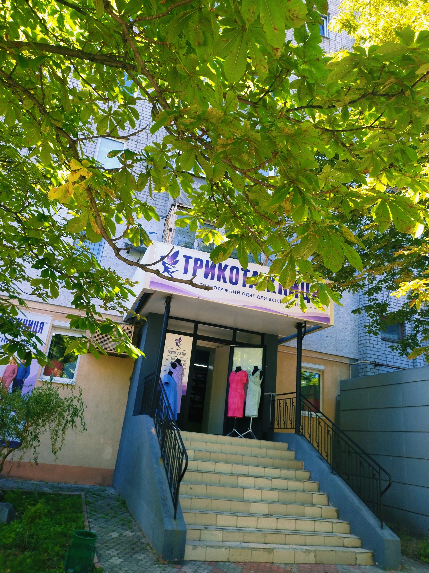 The image shows the entrance to a store called "TRIKOTAGE" in Horishni Plavni, Ukraine. The store is located in a building with a light-colored brick facade. A large tree with green leaves partially covers the storefront. The entrance features a set of stairs leading up to a covered area. Inside the store, mannequins display clothing. A sign above the entrance reads "TRIKOTAGE" in large letters, with smaller text below indicating it sells knitted clothing for the whole family. Another sign on the building to the left displays images of people wearing clothing. The exterior of the building is a mix of beige and light gray. To the right of the entrance, a modern gray wall is visible. Green bushes and a trash can are situated in front of the store. The scene appears to be a sunny day.