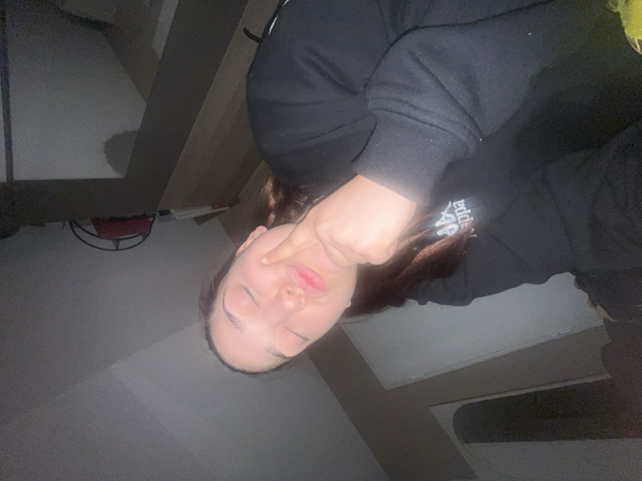 A young woman poses indoors, eyes closed and lips pursed, playfully touching her cheek with her index finger. She wears a black hooded sweatshirt with white lettering on the sleeve. The room is dimly lit, with a bed visible in the background. The image is rotated counterclockwise, creating a tilted perspective.