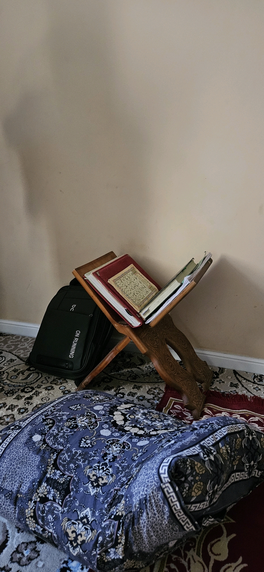 A medium shot captures an indoor scene with a prayer mat and religious texts. The foreground features a dark blue and white patterned rug, partially covering a larger, intricately designed carpet. Behind the rug, a dark green backpack with white lettering "ON RUNNING" rests against a wall. To the right of the backpack, a wooden book stand displays several books, including one open to pages with Arabic script. The background consists of a plain, light-colored wall. The lighting appears natural, casting soft shadows. The scene is devoid of people and any specific identifying features of Dushanbe, Tajikistan.