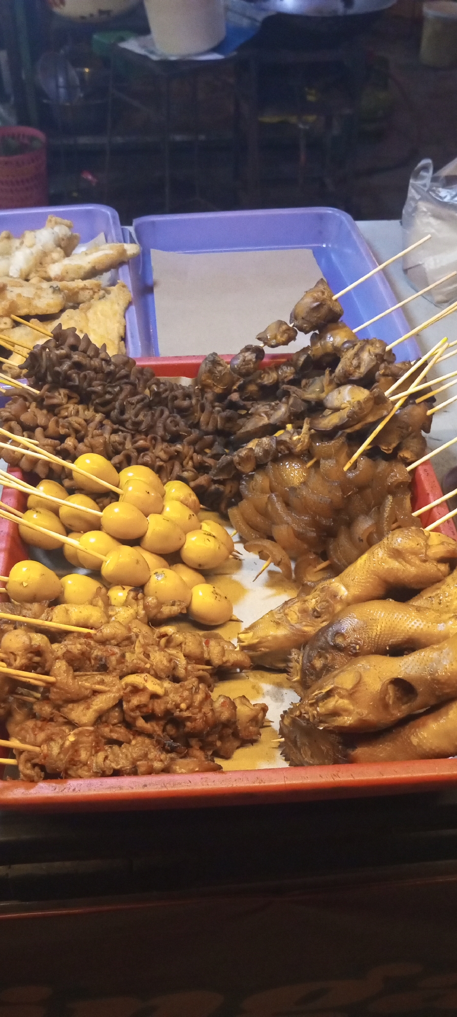 In a street food stall in Setu, Indonesia, a tray of various grilled skewers is displayed. The skewers contain what appear to be small, whole fish heads and necks, cut up intestines, quail eggs, and pieces of meat that could be chicken or pork. The food is arranged in different sections within the red tray, with bamboo skewers piercing the items. Some of the food is submerged in a light brown sauce. The stall appears to be outdoors, possibly at night, given the lighting. In the background, other food stalls and people can be seen blurred.