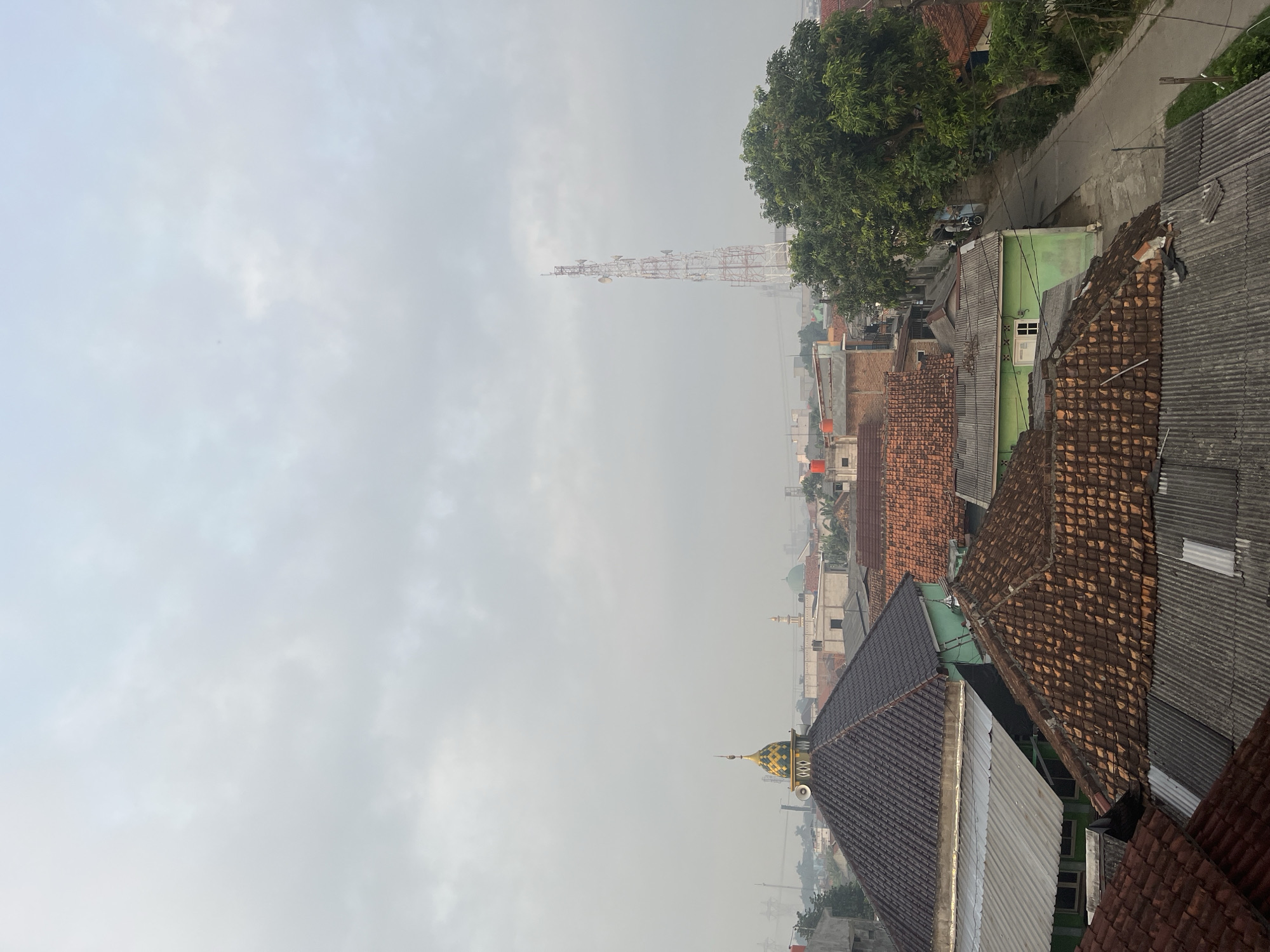 The image depicts a cityscape of Pasir Gadung, Indonesia, under a heavily overcast sky. The foreground is dominated by the roofs of residential buildings, characterized by varying materials such as clay tiles in shades of brown and grey corrugated metal. A prominent structure with a dark brown tiled roof features a distinctive gold and green domed top, adorned with a speaker or loudspeaker. Adjacent to this, another building exhibits a bright green facade.
In the midground, the urban landscape unfolds with more houses, some displaying orange tiled roofs. A tall, red and white lattice telecommunications tower stands prominently in the middle distance, equipped with multiple antennas. To the right of the tower, a large, leafy green tree partially obscures a paved road where a person on a bicycle is visible. Further in the background, several mosques are identifiable by their domes, including one with a green dome. The atmosphere suggests a hazy, possibly humid environment, with the distant buildings appearing softened by the atmospheric conditions. The overall scene captures a typical residential area within a developing urban setting in Indonesia.