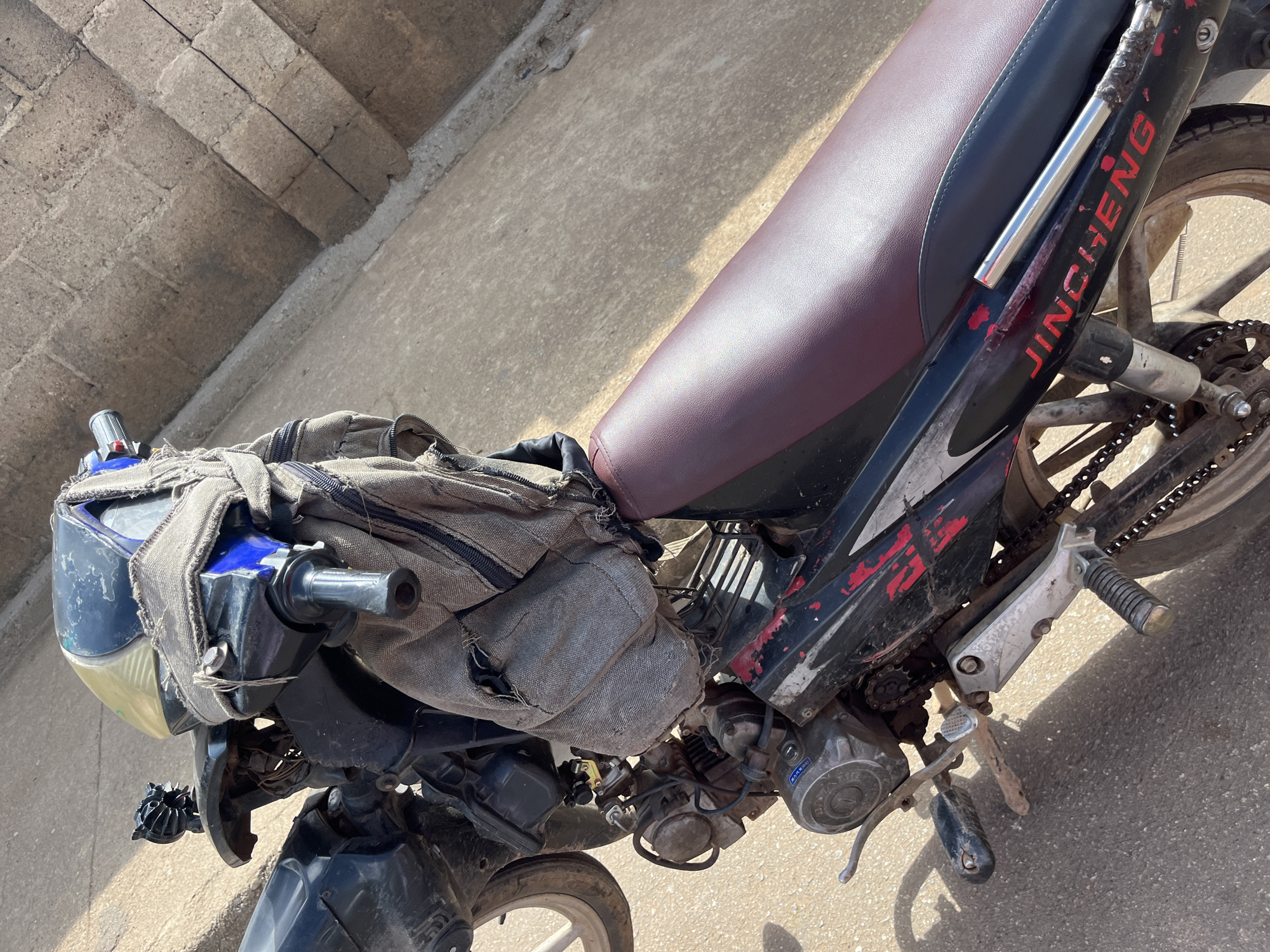 A medium shot captures a close-up view of a black and red motorcycle parked outdoors on a paved surface, possibly in Jos, Nigeria. The motorcycle is weathered and appears to have some makeshift repairs, including a rough, tan-colored fabric wrapped around the handlebars and part of the front. A portion of the motorcycle's engine and drivetrain are visible, with a chain and sprocket system clearly discernible. The seat of the motorcycle is a deep maroon color. A portion of a textured, brownish-gray wall or structure is visible in the upper right quadrant of the image. The overall lighting suggests it is daytime, with shadows cast on the ground.
