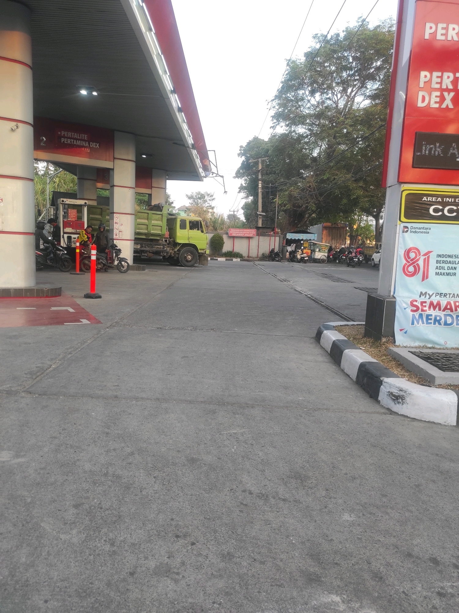 A view from a sidewalk looks across a paved area at a Pertamina gas station in Padalarang, Indonesia. The gas station has a covered canopy with fluorescent lighting. Underneath the canopy, a bright green dump truck is parked next to a gas pump. Several motorcycles are parked near the pump, and two people are standing beside them. A red and white sign with "PERTALITE PERTAMINA DEX" is visible on the canopy. To the right, a tall red and white sign displays "PERTAMINA" at the top, followed by "DEX". Below "DEX", a section reads "LINK A". Further down, a yellow sign with the words "AREA IN DI" and "CCTV" is present. Below that, a blue sign has a large red number "81" and text about Indonesia. At the very bottom of the red and white sign, the words "MyPERTAMINA SEMARAK MERDEKA" are visible in large letters. The ground is paved with asphalt. Trees and some buildings are visible in the background. The sky is overcast.