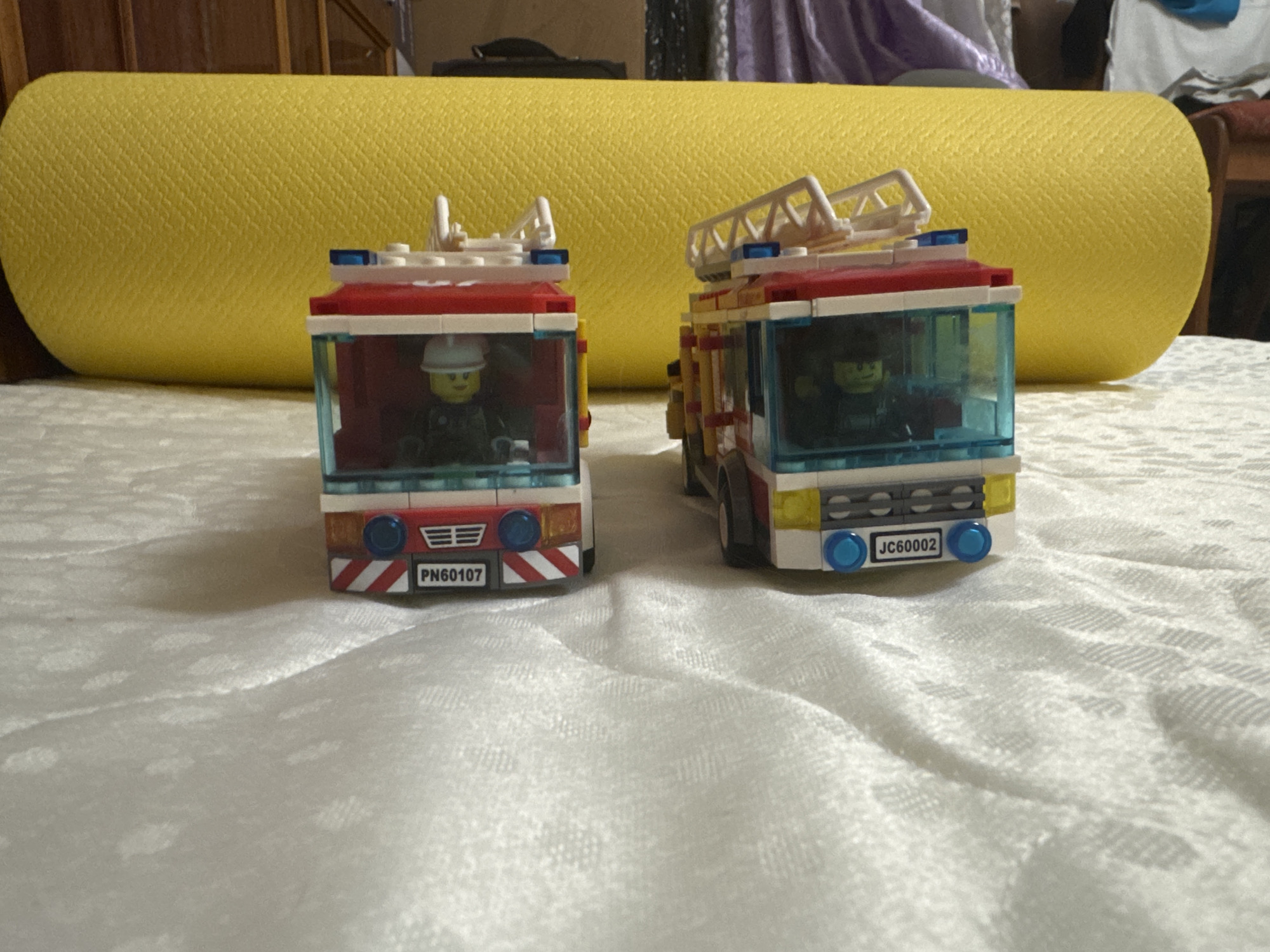 Two Lego fire trucks, each with a minifigure driver, are positioned side-by-side on a white, textured surface. The fire trucks are red and white with blue accents and ladders. The one on the left has a license plate reading "PN60107," and the one on the right has a license plate reading "JC60002." Behind the fire trucks is a large, bright yellow cylindrical object, possibly a yoga mat, which is positioned horizontally. The background is blurred, showing hints of wooden furniture and fabric. The scene appears to be indoors.