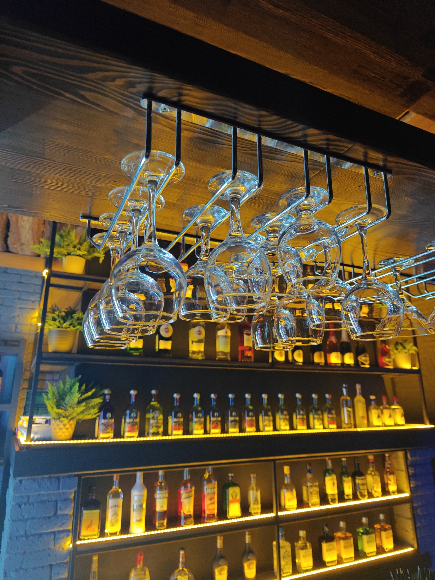 The image depicts the interior of a bar in Chaikovsky, Russia. A wooden ceiling supports a metal rack holding numerous empty wine glasses upside down. Below, shelves lined with various liquor bottles are illuminated by warm, built-in lighting. Potted green plants are visible on the upper shelf. The backdrop consists of a blue-toned brick wall.