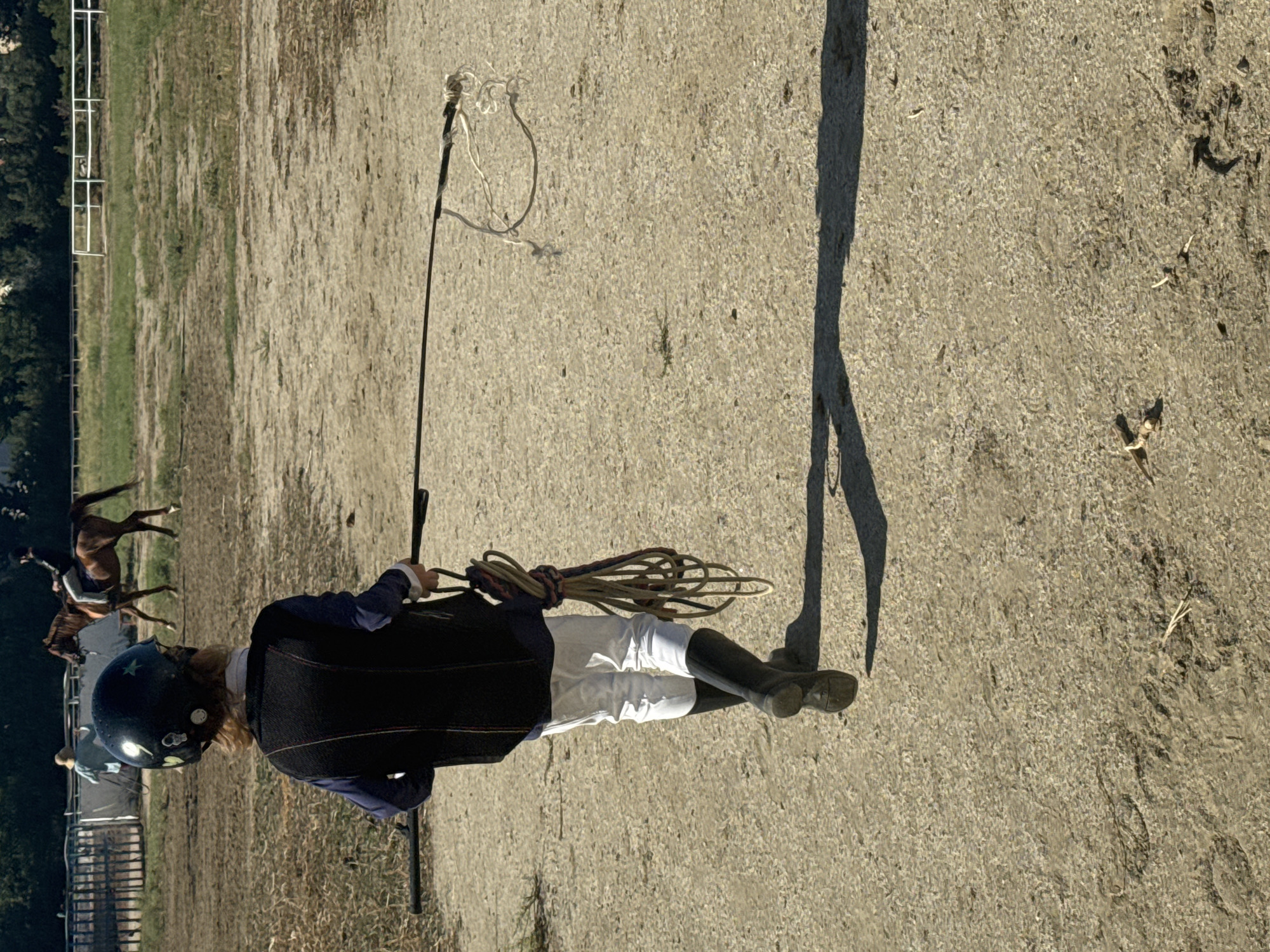 The image shows a young person wearing riding attire, including a black helmet, a dark vest over a blue shirt, white riding pants, and black boots. They are walking away from the camera, holding a riding crop in their right hand and a coiled rope in their left. In the background, two horses and riders are visible in a grassy field with a white fence. The ground in the foreground is a dirt or gravel surface. There are no discernible landmarks or features that place this scene in Baku, Azerbaijan.