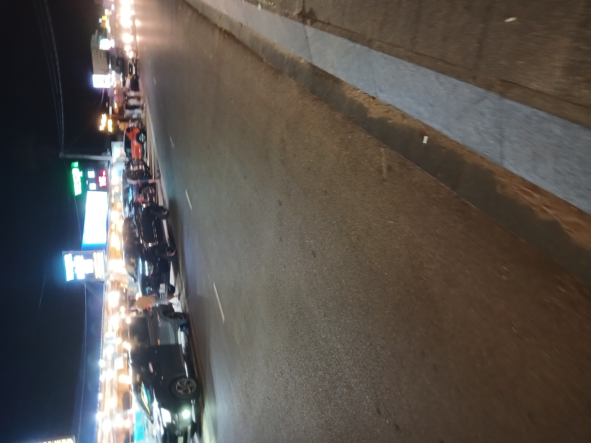The image depicts a street scene at night in Phu Thuy Ward, Vietnam. The street is paved and has a white line marking one lane. On the left side of the street, several vehicles are parked or moving, including a dark-colored pickup truck with its headlights on, and another dark-colored SUV. There are also people visible standing on the sidewalk and near the vehicles. Buildings with illuminated signs line the street on the left. The right side of the street features a sidewalk and a curb, with more vehicles and distant streetlights visible further down the road. The overall impression is of a busy commercial area at night.
