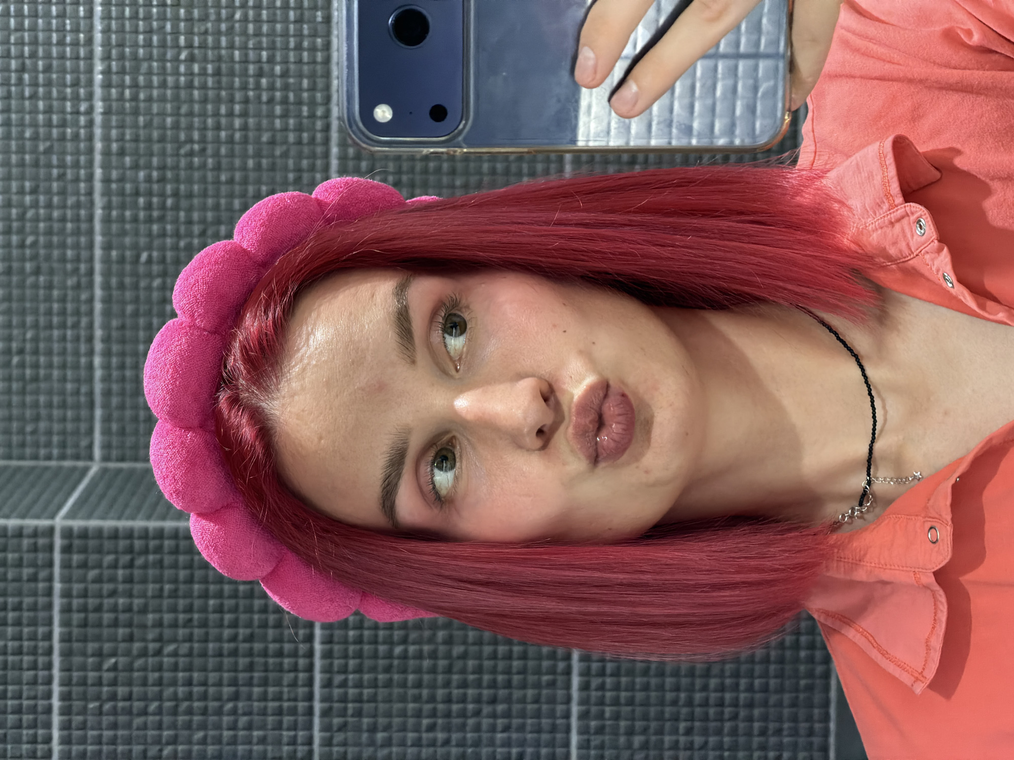The image depicts a young woman in an indoor setting within the city of Chaikovsky, Russia. She has bright pink, shoulder-length hair and is wearing a textured, plush pink headband composed of interconnected spherical segments. Her complexion is light, with visible blush on her cheeks and subtle eye makeup. Her lips are pursed into a pucker, and her eyes are looking to her left (viewer's right). She wears a coral-colored collared shirt with snap buttons and a black choker necklace adorned with a small silver star charm and a short chain segment. She is taking a selfie, holding a dark blue or silver smartphone with a reflective case in her right hand, partially obscuring the lower right portion of the frame. The phone's camera module, including multiple lenses and a flash, is clearly visible. The background consists of a dark gray wall covered in small, raised square tiles, arranged in a grid pattern. The lighting is bright and even.