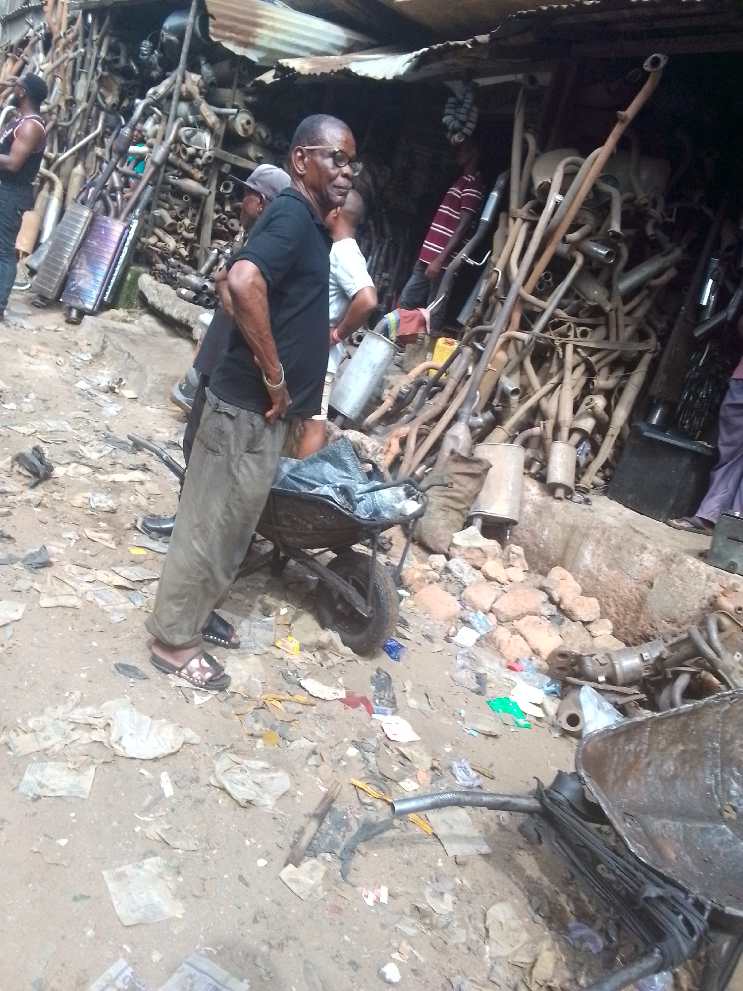 This media file captures an outdoor auto spare parts market or junkyard in Onitsha, Nigeria, during daylight hours. The scene is dominated by large, haphazard piles of metallic vehicle components, primarily exhaust pipes and mufflers, some appearing rusty and used, others shinier. These components are stacked high, forming walls or displays under a corrugated metal roof, typical of informal commercial settings.
In the foreground, a Black man with glasses, wearing a black short-sleeved shirt, olive green cargo pants, and dark sandals, stands facing right with his hands behind his back. A gold-colored bangle is visible on his right wrist. Text on his cargo pocket includes "COLLECTION" and "JEANS," with "BOSS" or "DOSS" above. Behind him, another man in a light shirt is partially visible. To the left, a man in a black tank top looks towards the metal stacks. Further back, another individual in a red and white striped shirt is seen among the parts.
The ground is unpaved, consisting of dirt and sand, and is heavily littered with various debris including torn papers, plastic bags, and small rocks. A black metal wheelbarrow is visible in the lower center, and a red jerrycan sits among the metal piles in the background. The overall impression is one of a bustling, informal market dealing in used auto parts. The people appear to be standing and observing, suggesting they are either customers, workers, or bystanders in this environment. There are no strong emotional expressions visible.