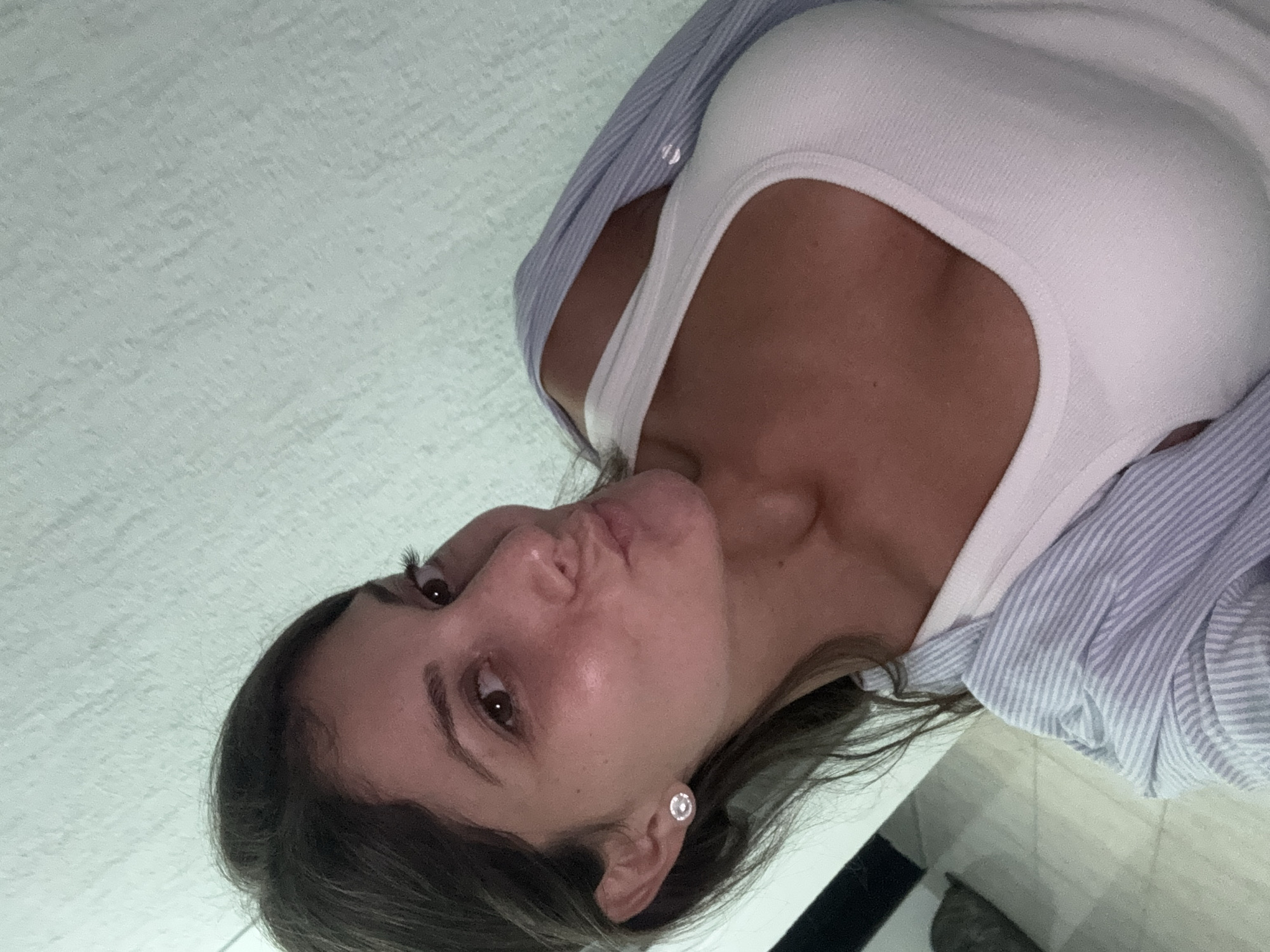 A woman is taking a selfie with a phone, her face angled towards the camera. She is wearing a white tank top and a blue and white striped open button-down shirt. Her hair is dark brown and pulled back, and she is wearing a stud earring on her left ear. The background appears to be an indoor setting with a textured light-colored wall. There are no visible landmarks or elements to suggest a specific location within Burgas, Bulgaria.