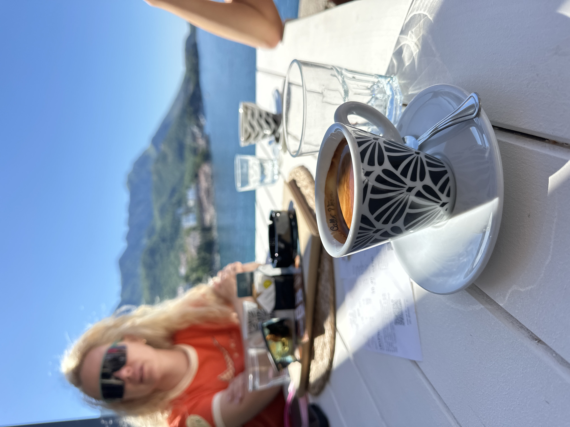 The image depicts a sunlit outdoor scene in Boreti, Montenegro, with a white wooden table in the foreground. On the table, there is a cup of coffee with a black and white patterned exterior on a white saucer with a spoon resting on it. Next to the cup are two empty glasses. A woman with long blonde hair and sunglasses is partially visible, looking at a cell phone. Other objects, including a hat, a black case, and a paper receipt are placed around her. In the blurred background, mountains, the sea, and buildings characteristic of the area are visible under a clear blue sky. A partially visible arm of another person is in the upper right corner.