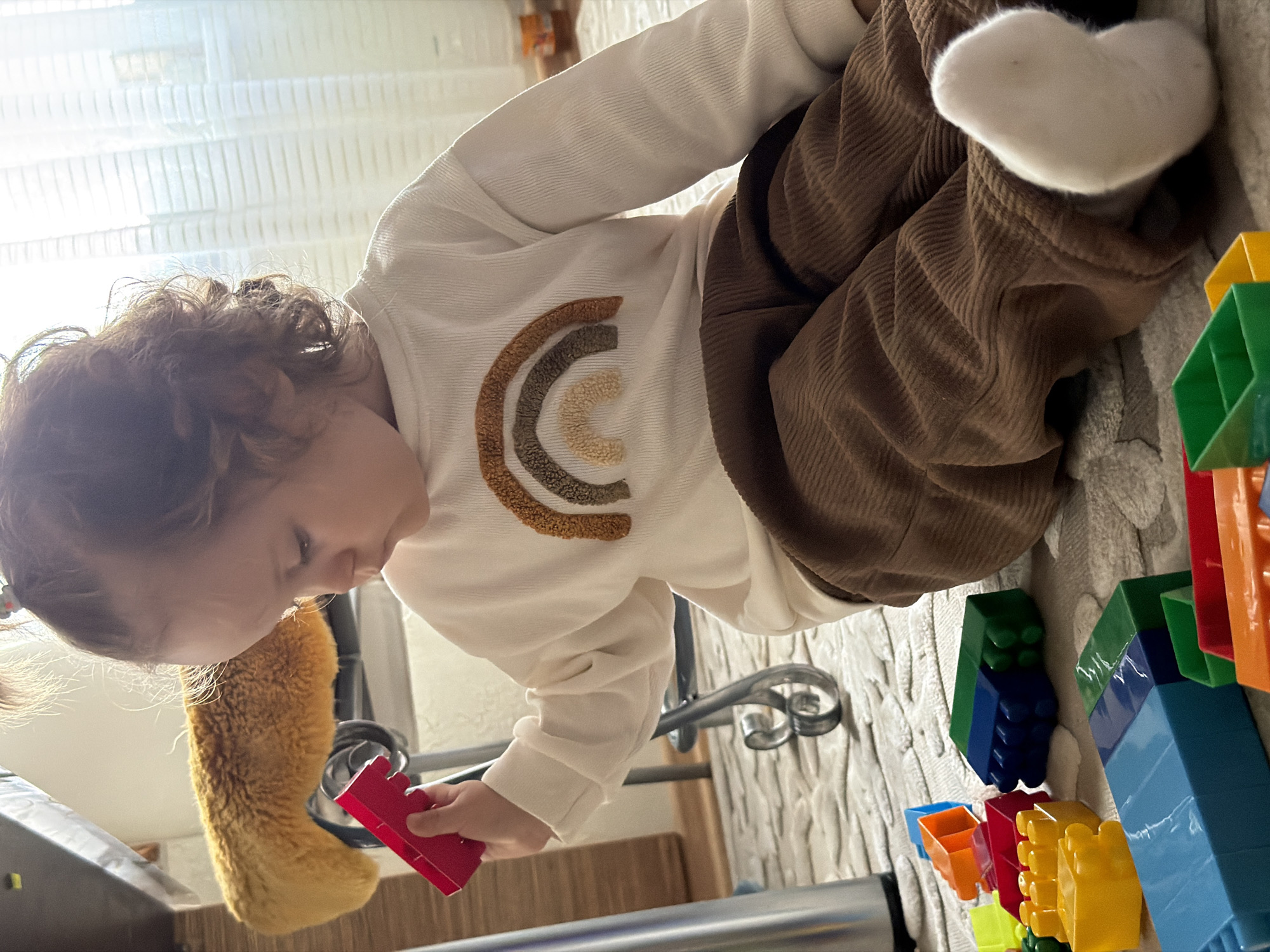 A young child with curly brown hair is seated on a rug, playing with building blocks. The child is wearing a white sweater with a brown and beige rainbow design and brown corduroy pants. The rug appears to be cable knit and off-white in color. There are various colored building blocks on the rug including green, blue, yellow, red, and orange. The child is holding a red block in their hand. There is a stair railing in the background along with a fuzzy, brown dog toy. The location cannot be determined to be Chaikovsky, Russia based on the provided image alone.