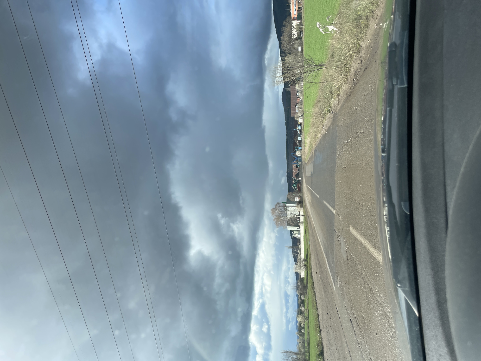 The image depicts a road running through a rural area. A row of houses and greenery line one side of the road, while power lines stretch across a cloudy sky on the other side. The photograph was taken from inside a vehicle. The exact location within Sofia, Bulgaria is not discernible from the image.