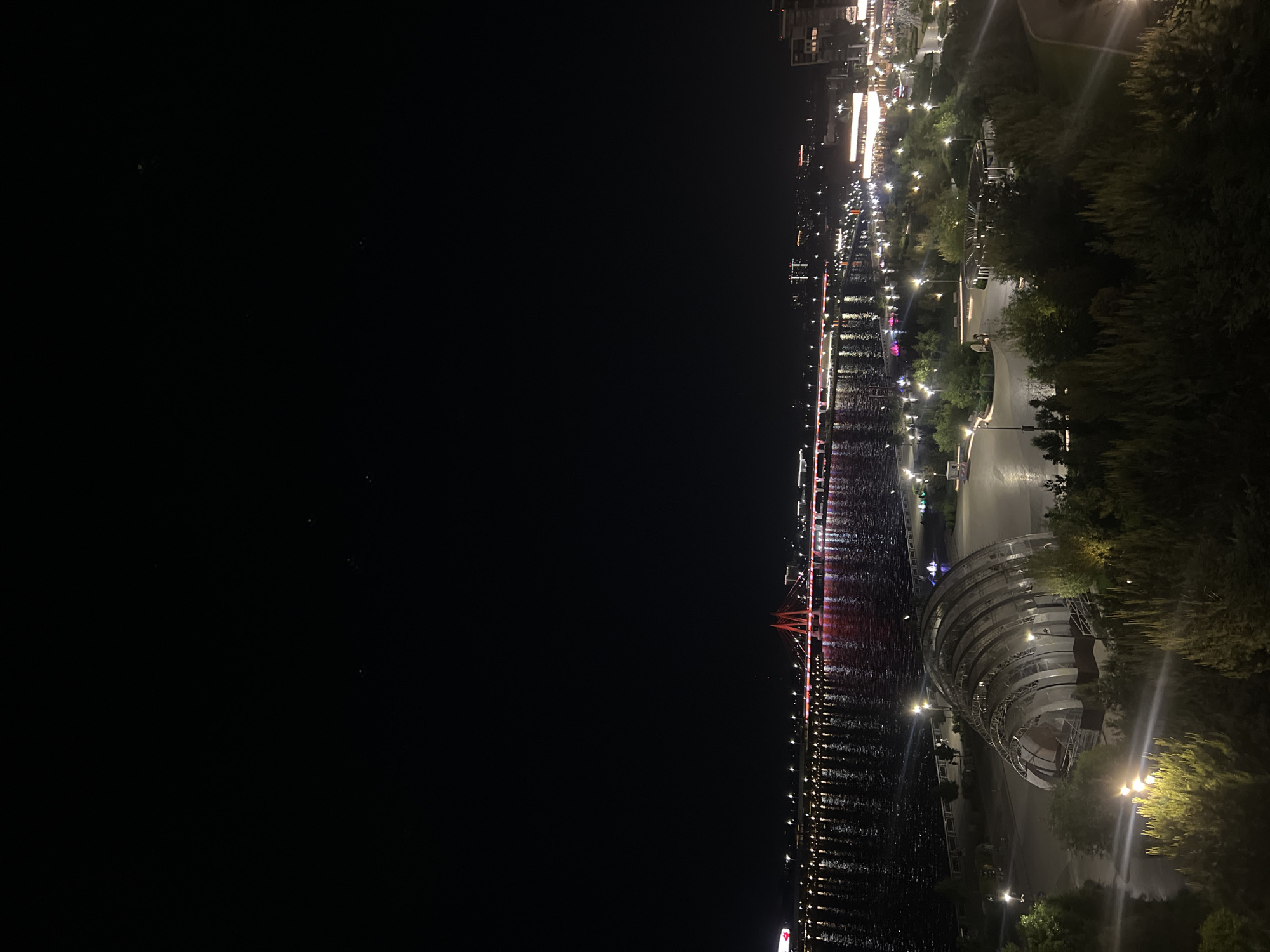 The image depicts a nighttime view of the city of Kazan, Russia, alongside the Kazanka River.
The right side of the frame showcases a portion of the city with illuminated buildings, trees, and walkways. A distinctive architectural structure, potentially a pavilion or event space, is situated near the waterfront. Various lights dot the scene, reflecting on the river's surface.
The left side of the image is predominantly dark, representing the night sky and the wider expanse of the river. The reflections of city lights appear as shimmering lines across the water. A bridge with red illumination is visible.