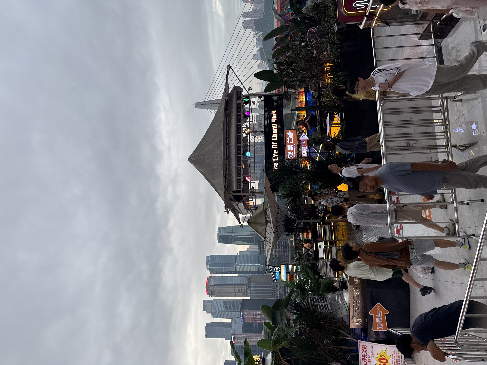 This image captures a view of "The Eye of Chongqing," a Ferris wheel attraction, situated in Yuzhong District, China. The scene is set against a backdrop of a cloudy sky and a dense urban skyline featuring modern skyscrapers. In the foreground, several people are visible, some walking, others standing. There is a raised platform with a covered structure, likely part of the attraction's amenities, which includes a bar area with bottles and lights, and what appears to be a stage with musical instruments. Plants and foliage are interspersed throughout the scene, adding a touch of greenery. Metal railings and barriers are present, indicating a controlled access area. A sign with Chinese characters and the words "Viewing Deck" is prominently displayed. The overall atmosphere suggests a public recreational space in an urban environment.