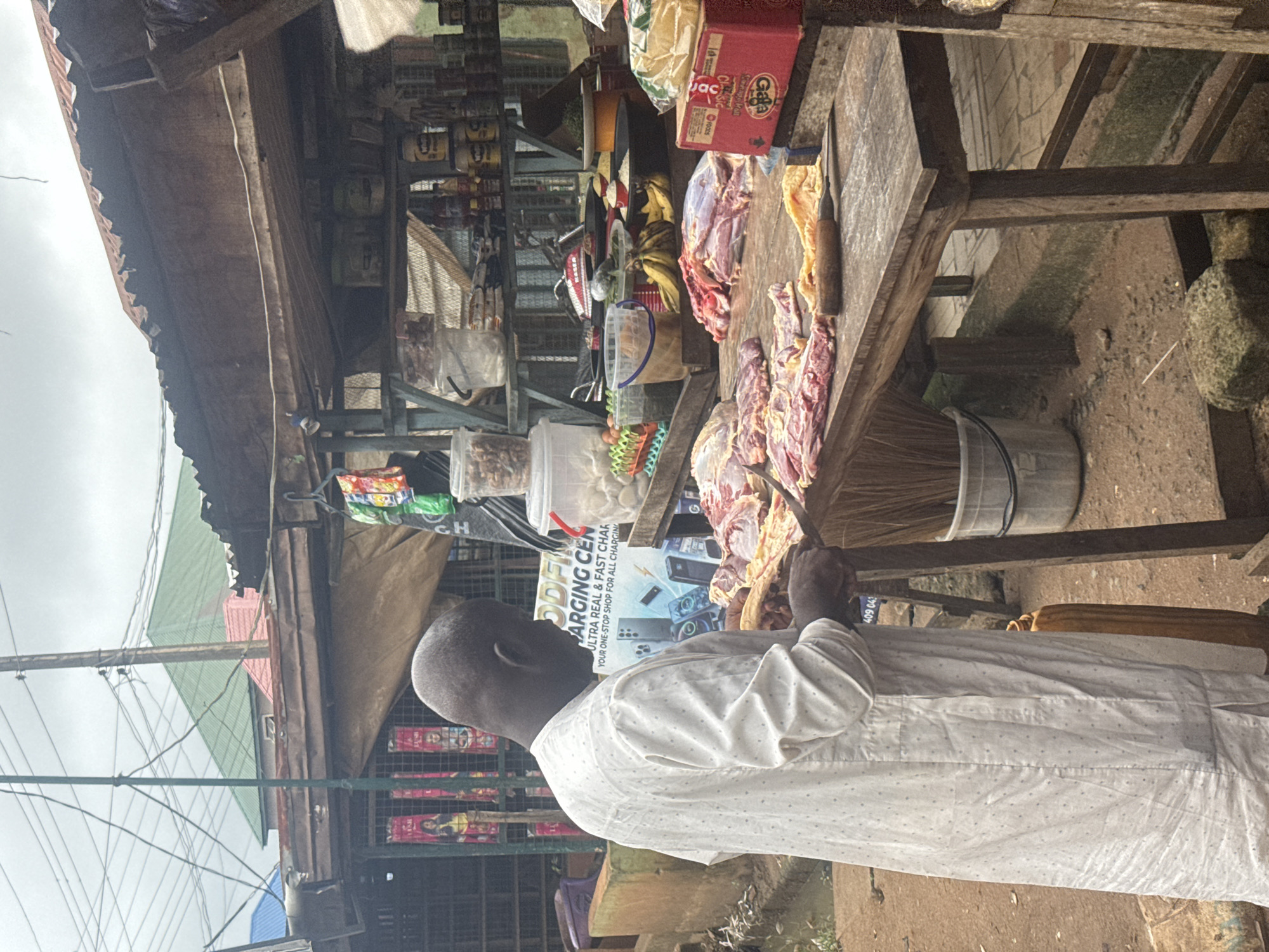 A man, wearing a white patterned robe, is seen from the side, standing at a wooden butcher's table in an outdoor market. He appears to be a butcher as he is in the process of cutting meat with a knife. The table is laden with raw cuts of meat. Behind the butcher's table, shelves are stocked with various goods, including canned items and packaged food. In the background, a market stall with a corrugated metal roof is visible, with various posters and bags hanging. The overall environment suggests a busy, functional marketplace in Ihiagwa, Nigeria. The ground is uneven and appears to be dirt or packed earth. Overhead power lines are also visible.