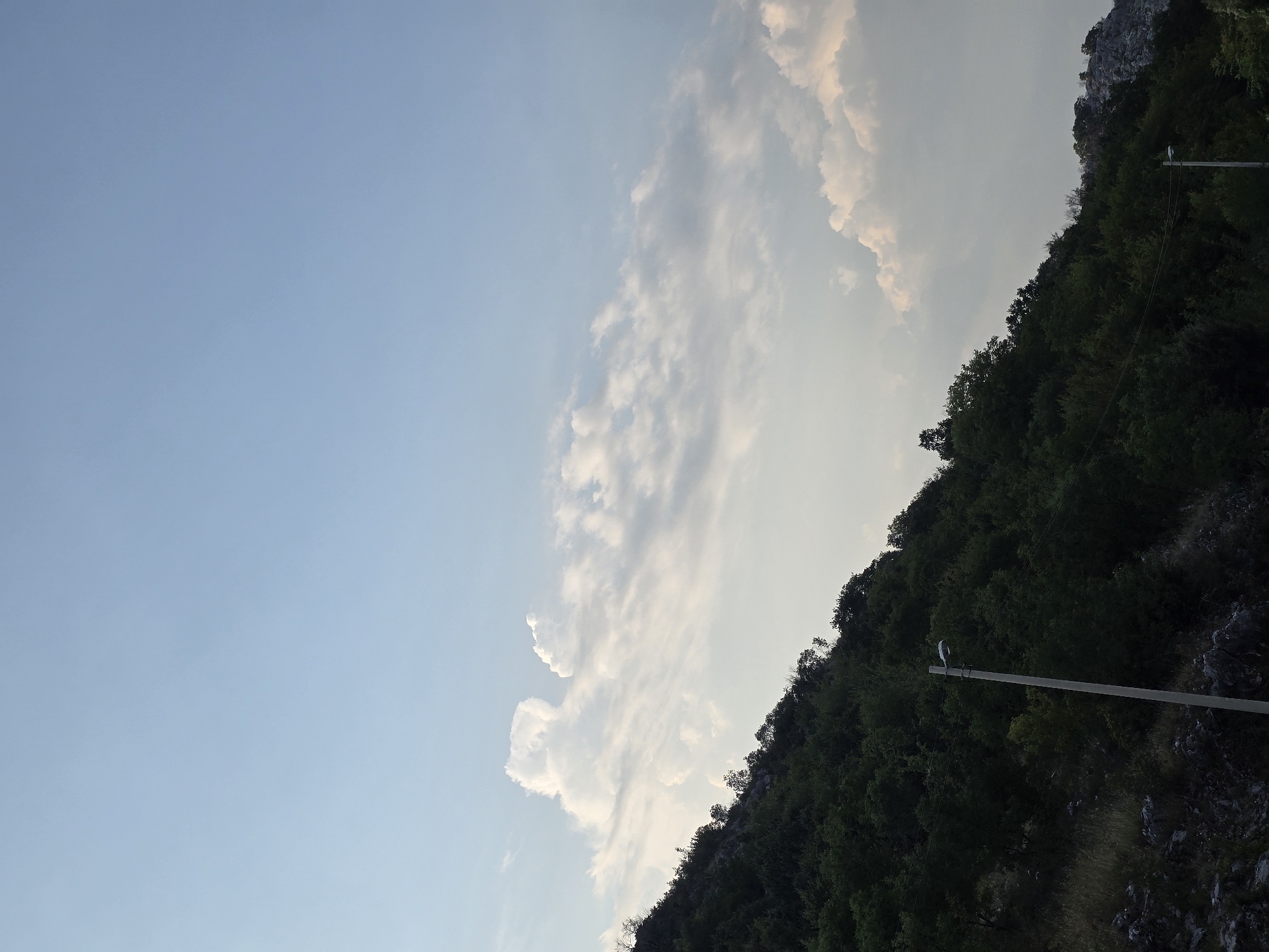 The image depicts a hillside covered in dense, dark green foliage, with rocky outcrops visible at the base. A single, tall, grey lamppost stands prominently in the foreground, extending upwards into the frame. Above the hillside, a vast expanse of pale blue sky dominates the upper portion of the image, with scattered, puffy white clouds. The lighting suggests it is either early morning or late afternoon. The location is identified as Orljevo, Montenegro.
